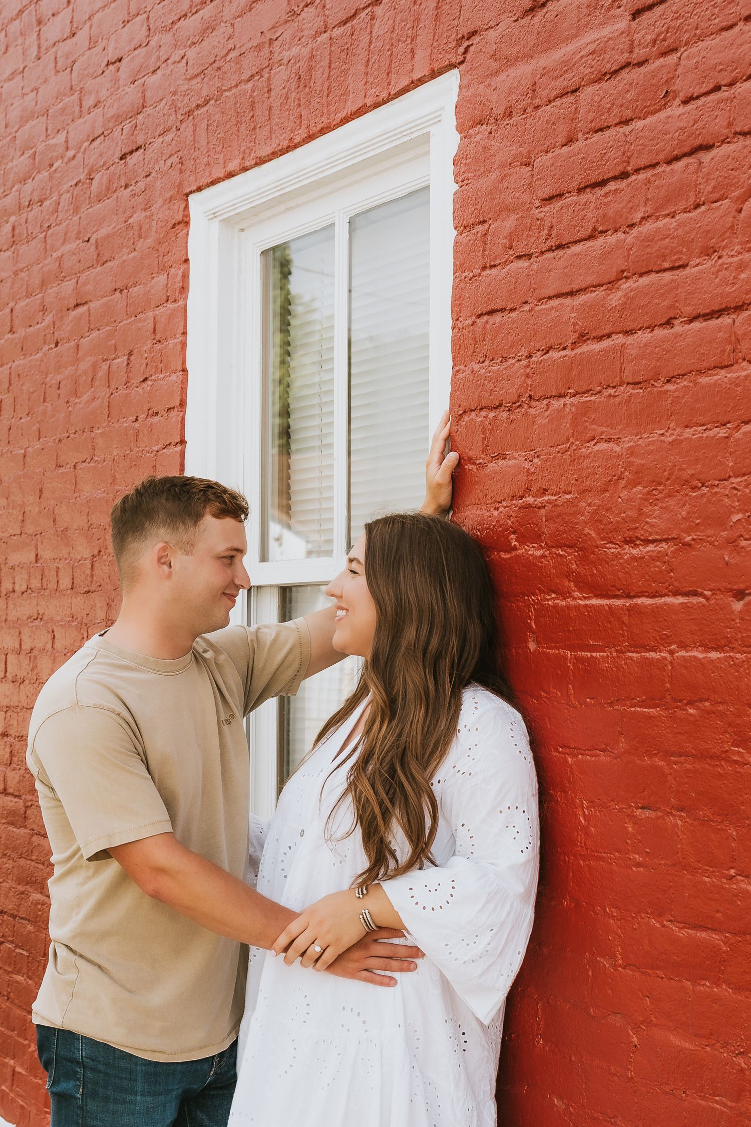 Couple posing against a red brick wall near a white window, man leaning over smiling woman in white dress.