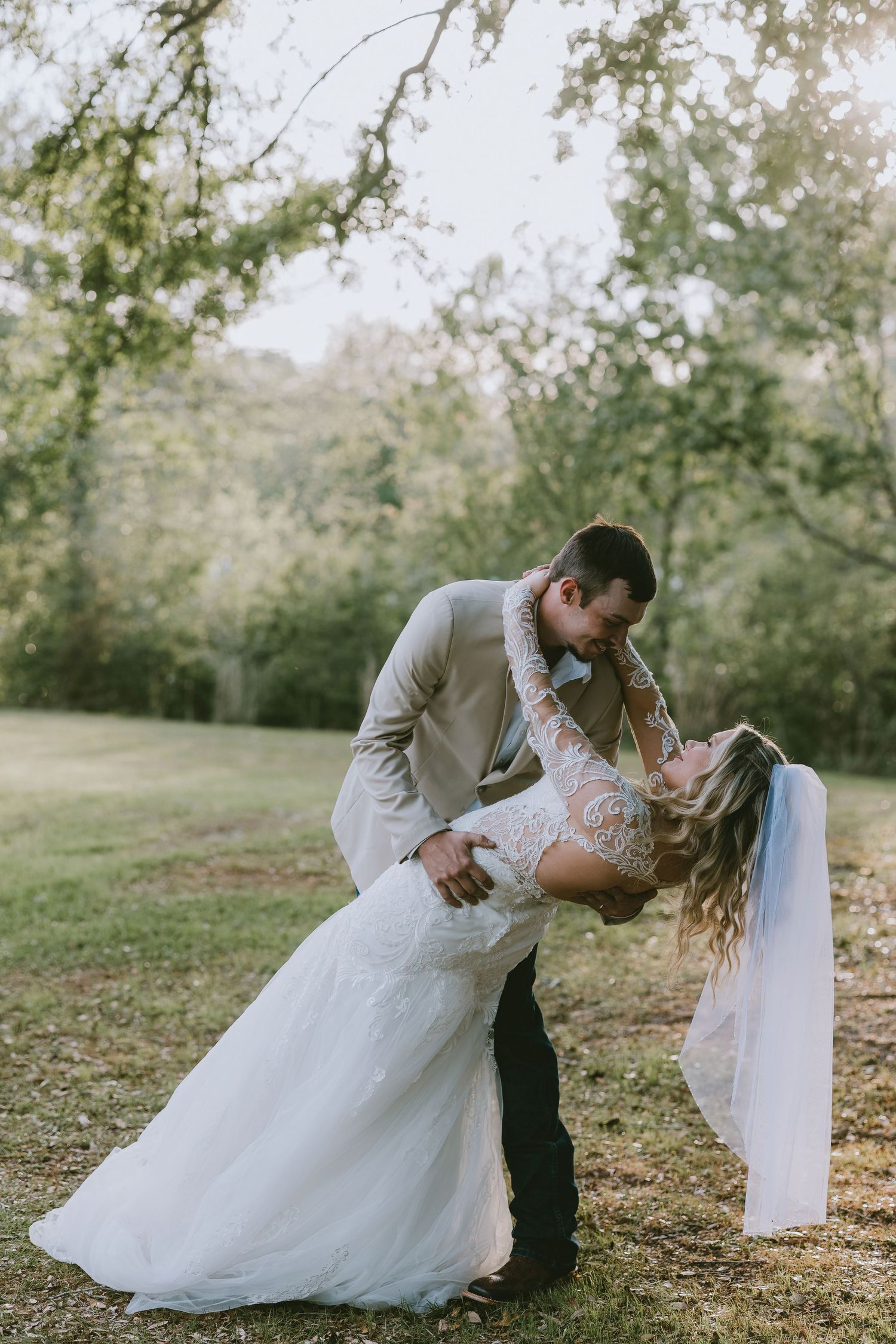 Bride and groom share a romantic dip kiss outdoors, surrounded by lush green trees on their wedding day.