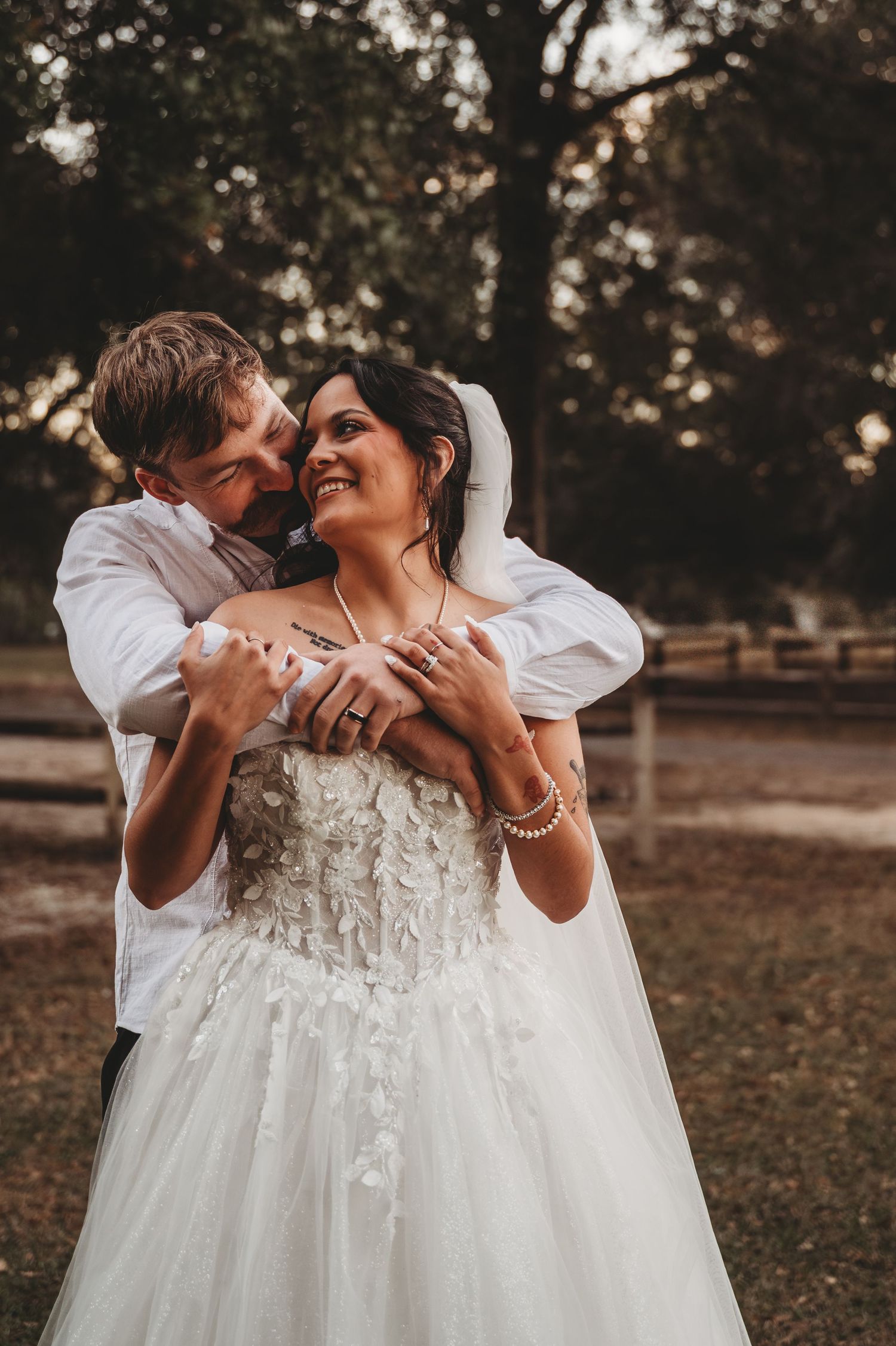 Groom embracing bride in floral strapless wedding gown and veil, smiling together outdoors near rustic fence.