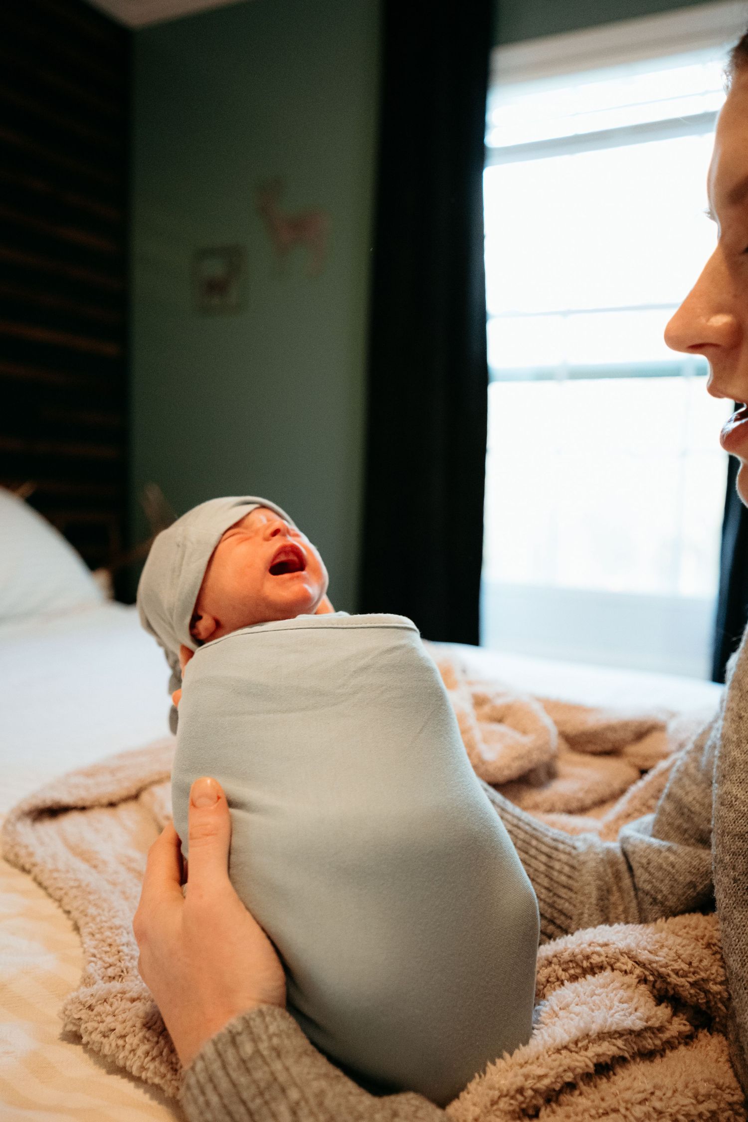 Mother holding a swaddled crying newborn baby in a light blue wrap with a knotted hat.