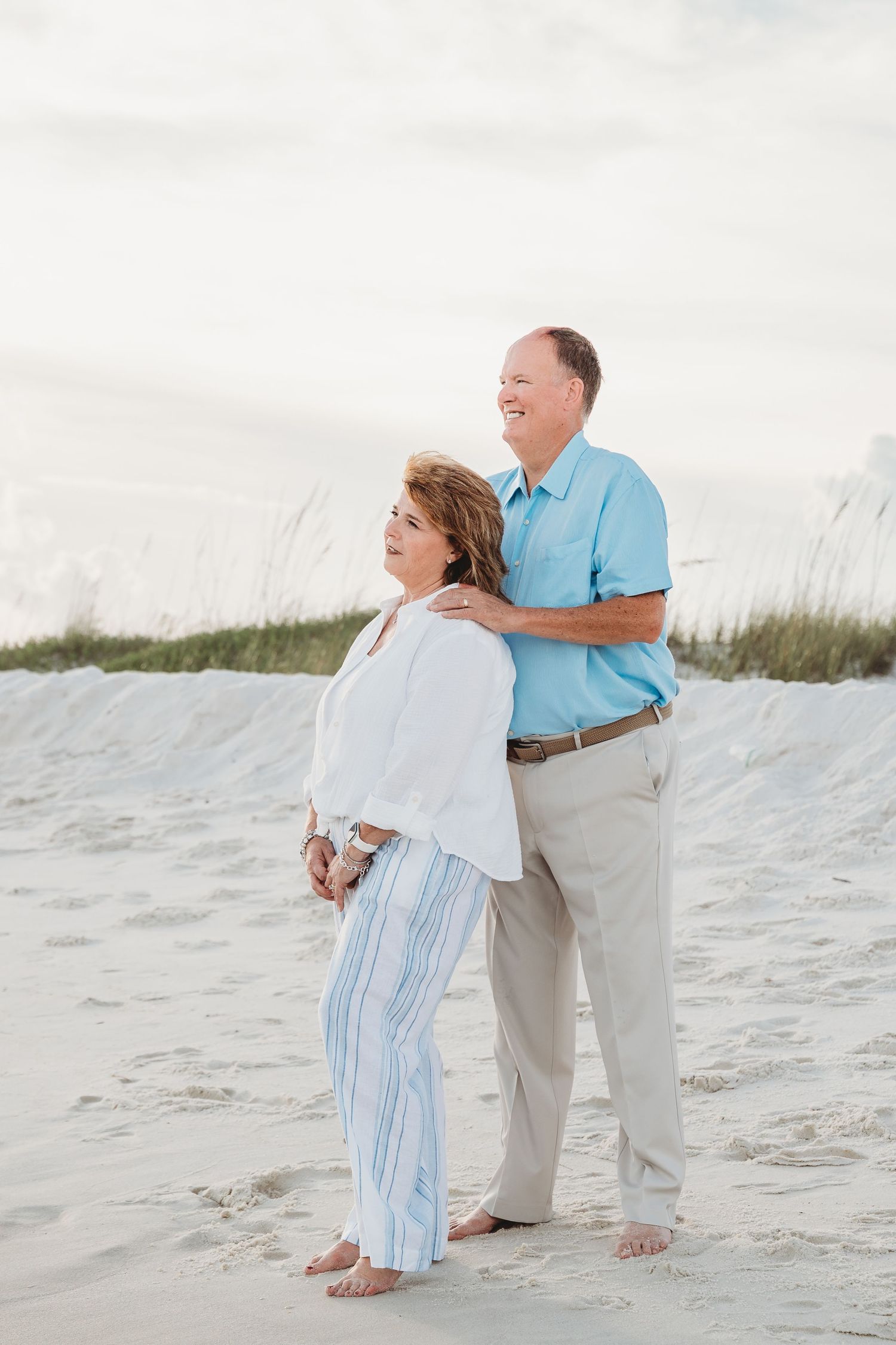 Couple standing together on white sand beach, man in blue polo embracing woman in white top and striped pants.