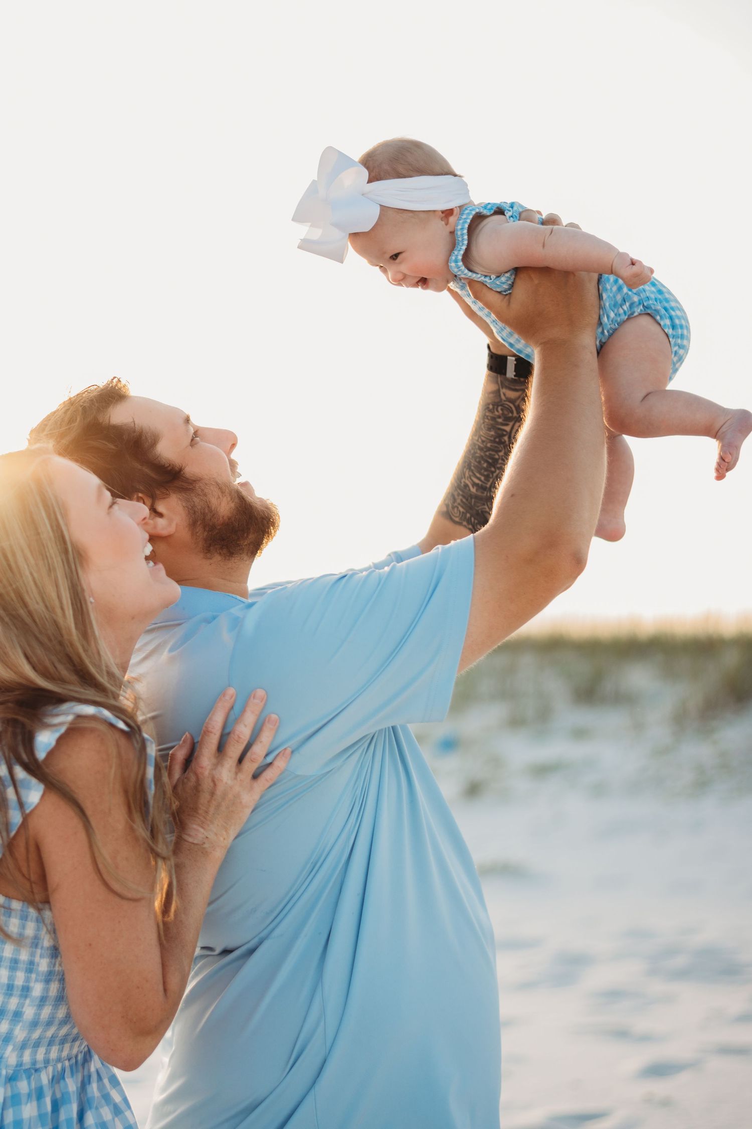 Parents lifting their baby girl on a sunny beach, all dressed in coordinating blue outfits.