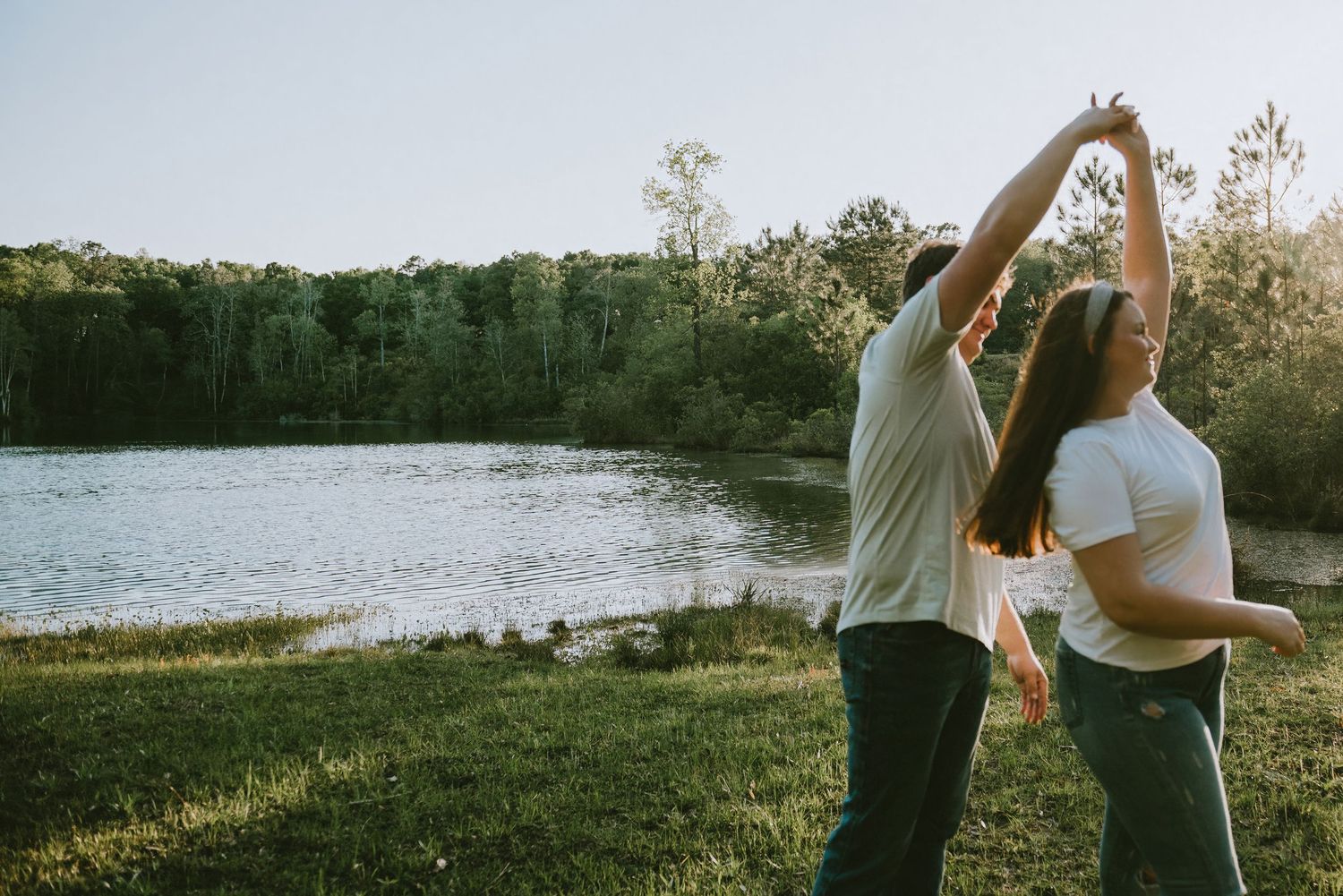 Couple dancing joyfully by a peaceful lake surrounded by lush green trees at sunset.