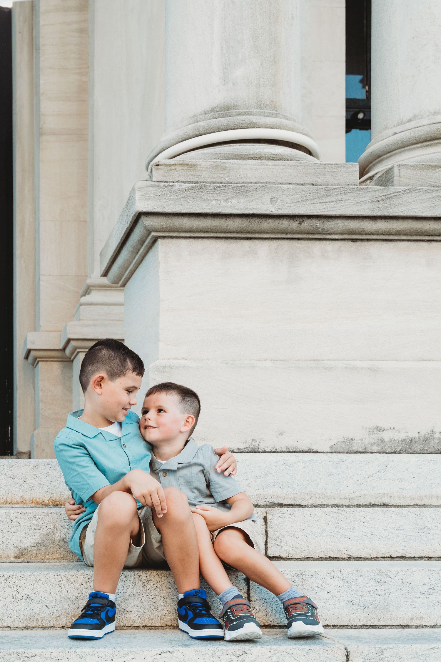 Two young brothers sit together on stone steps, sharing a tender embrace near a classical building.