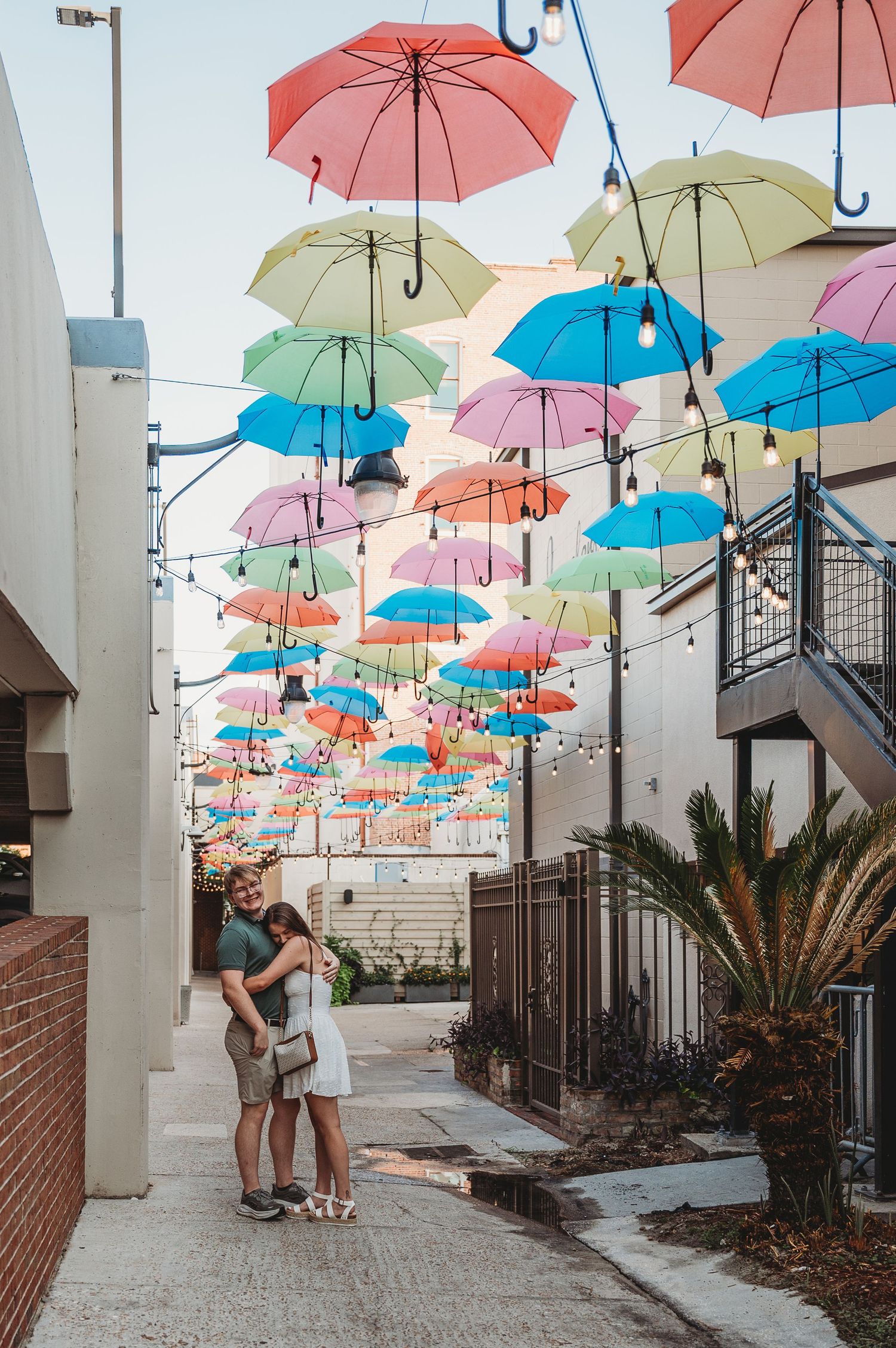 Couple embracing under colorful hanging umbrellas in a charming outdoor alleyway with string lights.