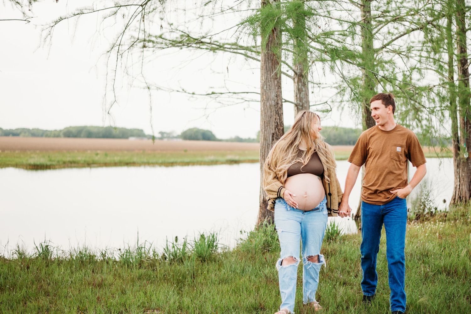 Expecting couple holds hands by a serene lakeside, showing off baby bump in a maternity photo.
