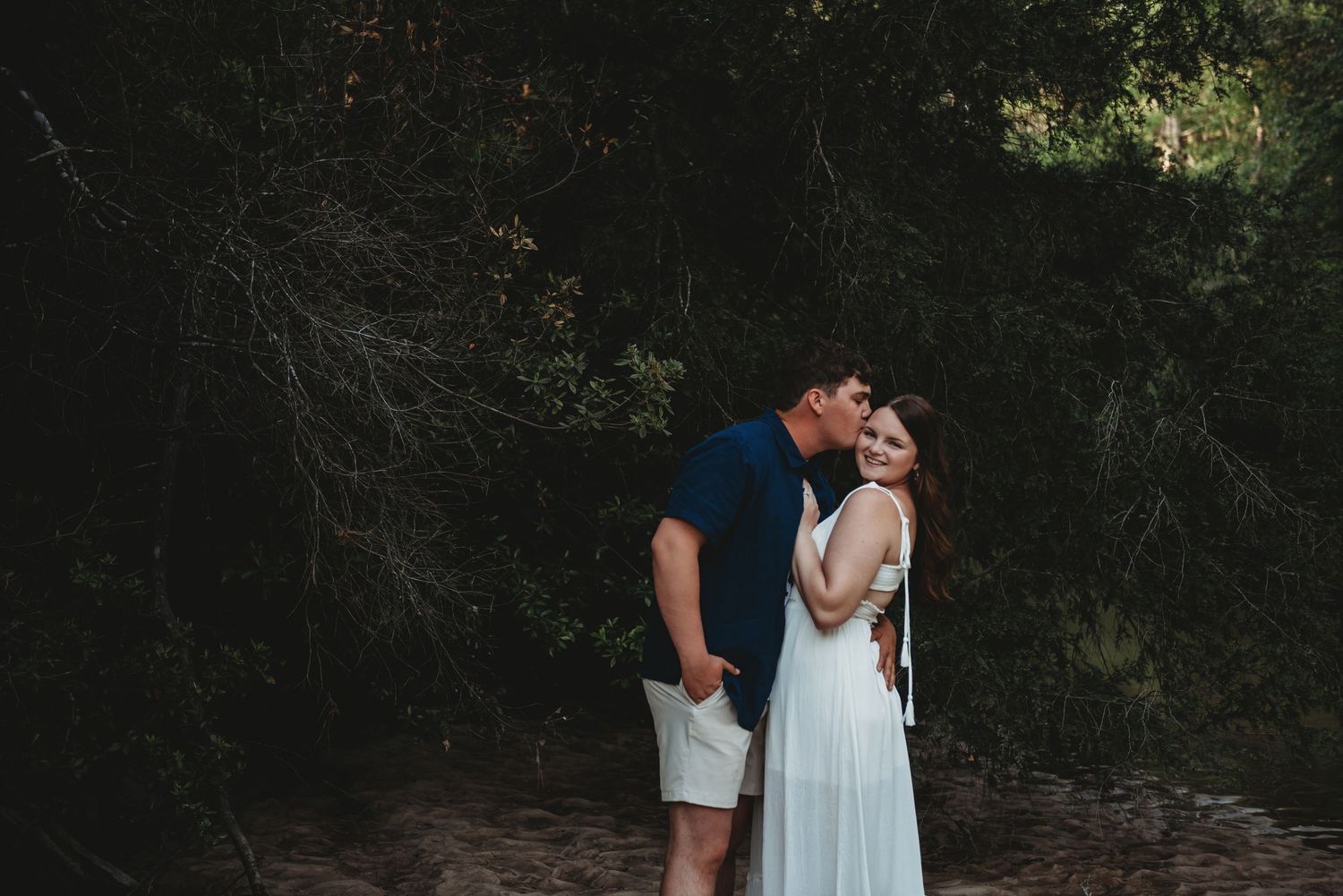 Man kissing his partner on the cheek as she smiles during an outdoor engagement photo session.
