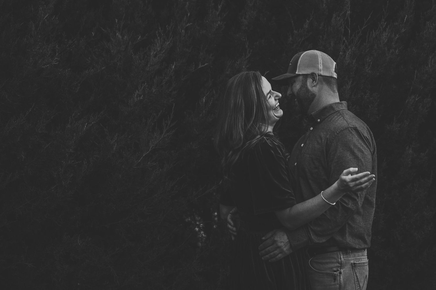 Black and white photo of a couple embracing and smiling at each other outdoors against a dark background.