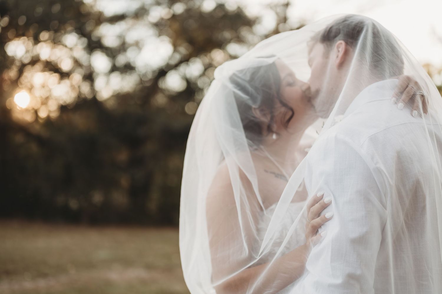 A bride and groom share a romantic kiss under a flowing white veil at golden hour outdoors.