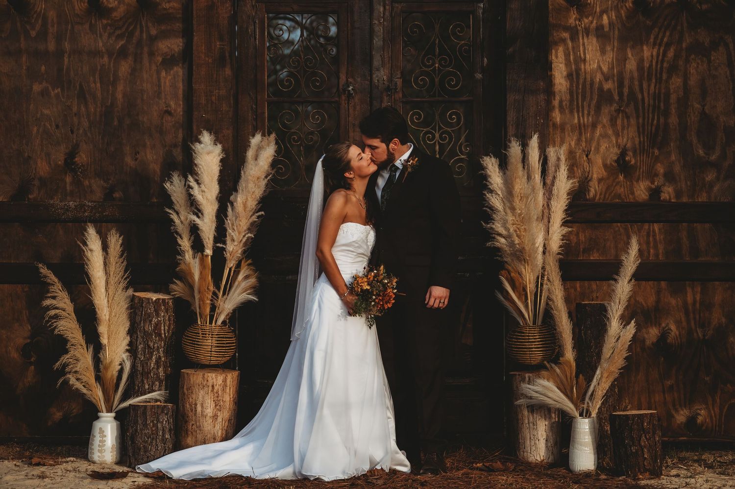 Bride and groom share a romantic kiss in rustic barn wedding venue with pampas grass decor and wooden backdrop.