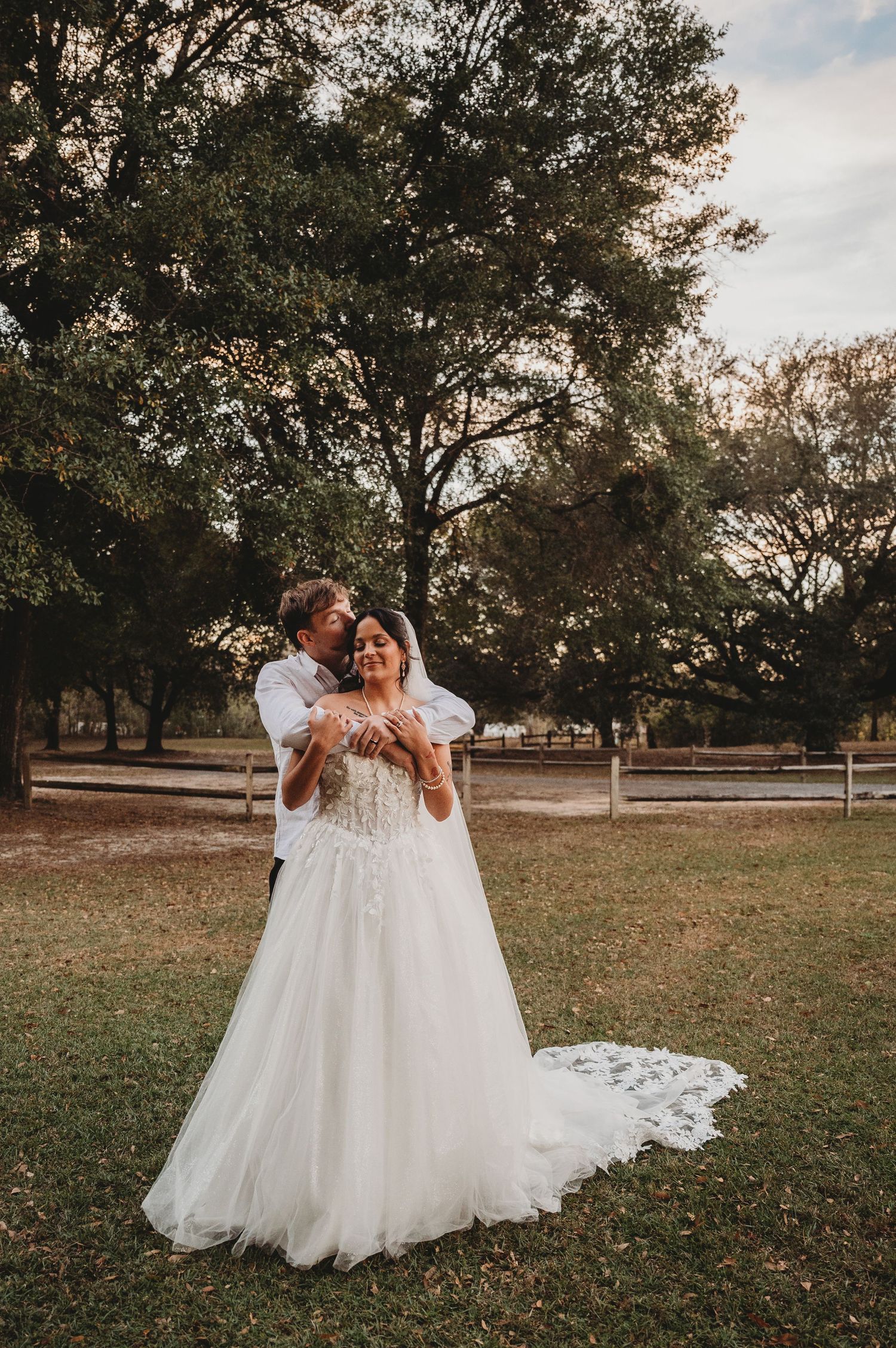 Bride in white ball gown with groom embracing her from behind in a lush outdoor wedding setting.