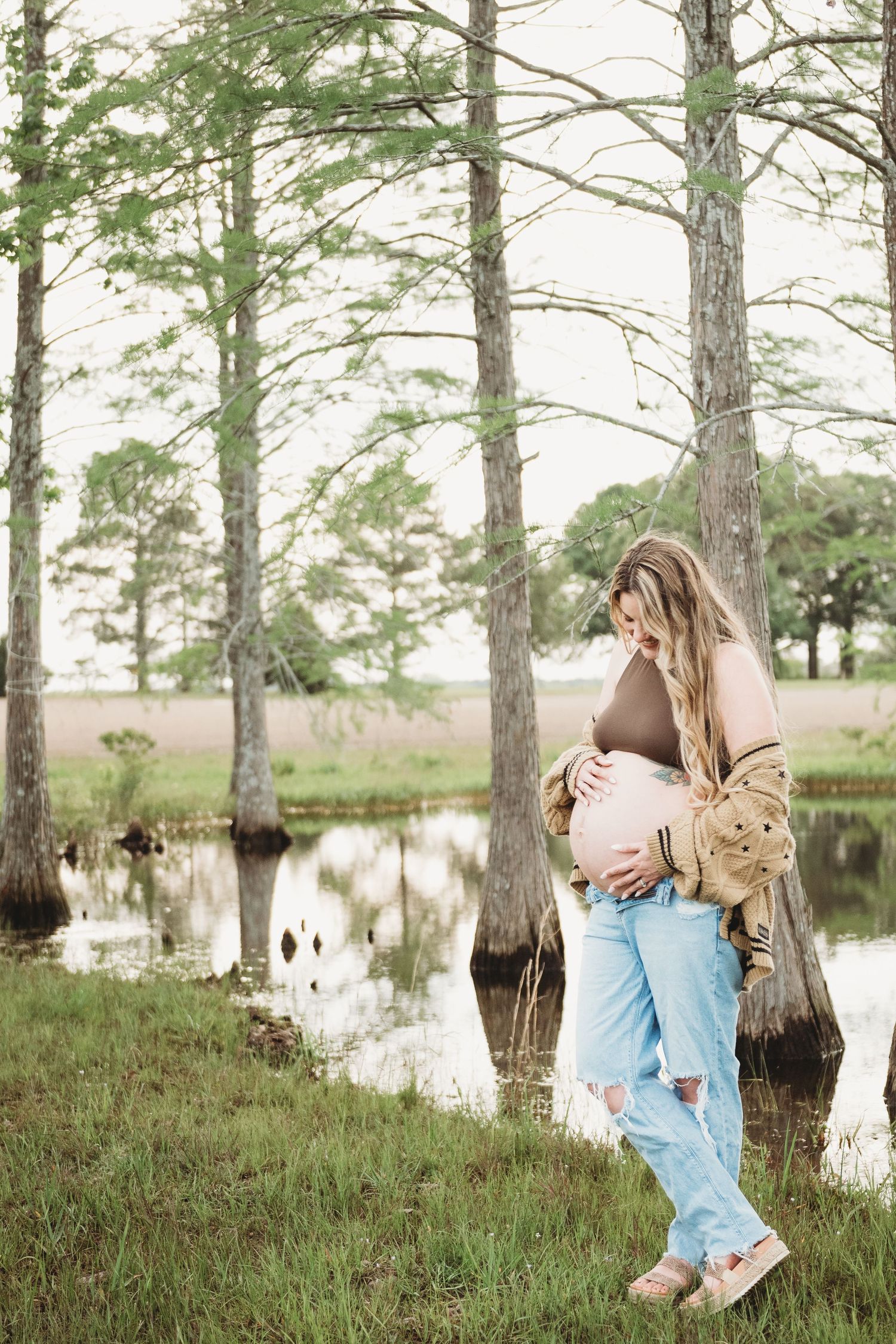 Pregnant woman in ripped jeans and brown top cradling baby bump near cypress trees by a tranquil pond.