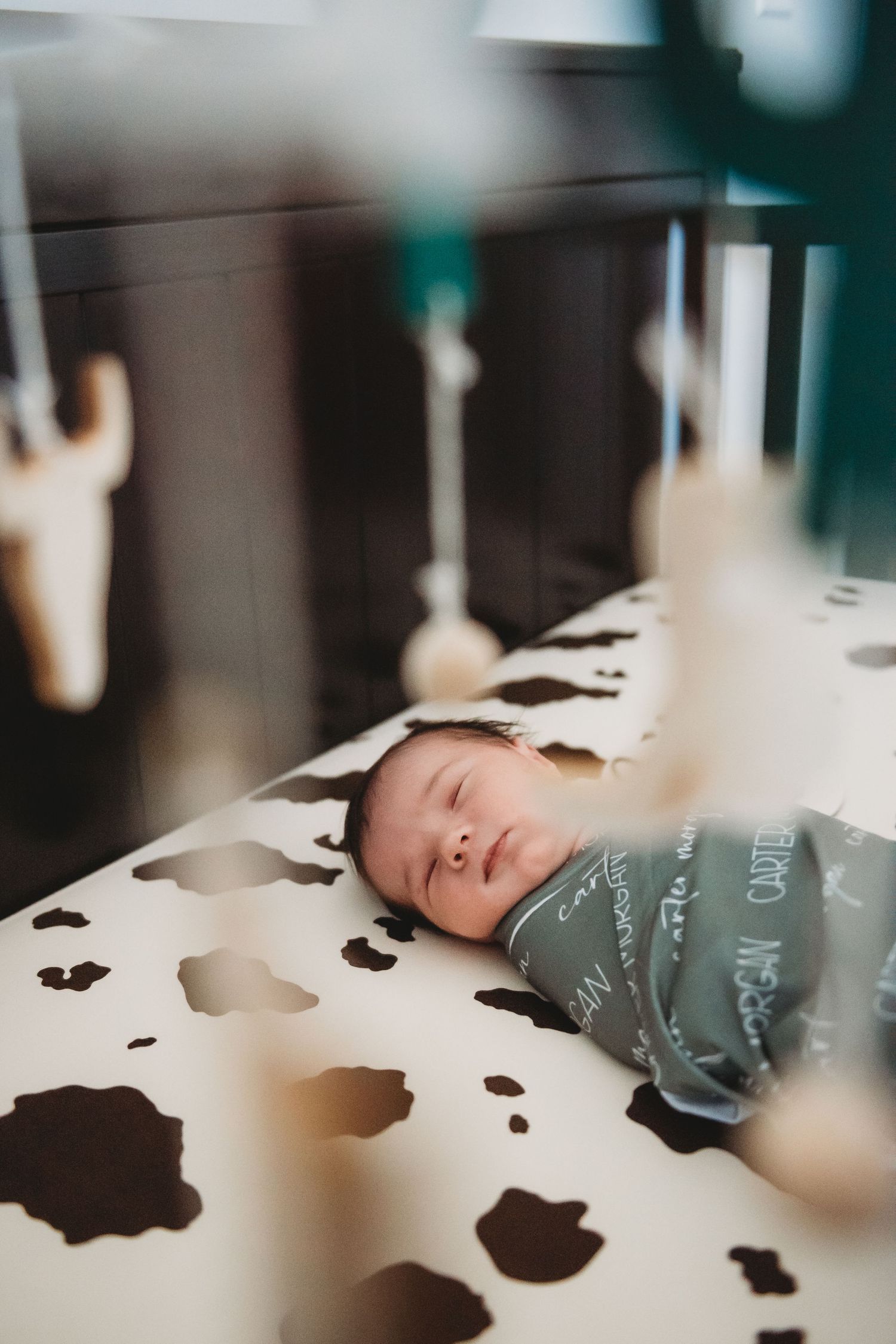 Swaddled newborn sleeping peacefully in a cow-print crib with a personalized name blanket.