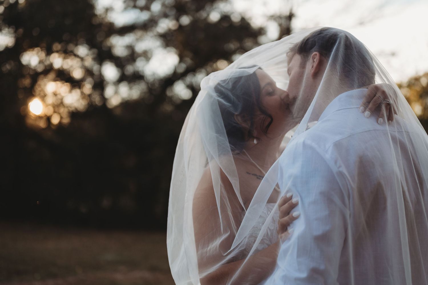 Bride and groom sharing a romantic kiss under a flowing wedding veil at golden hour outdoors.