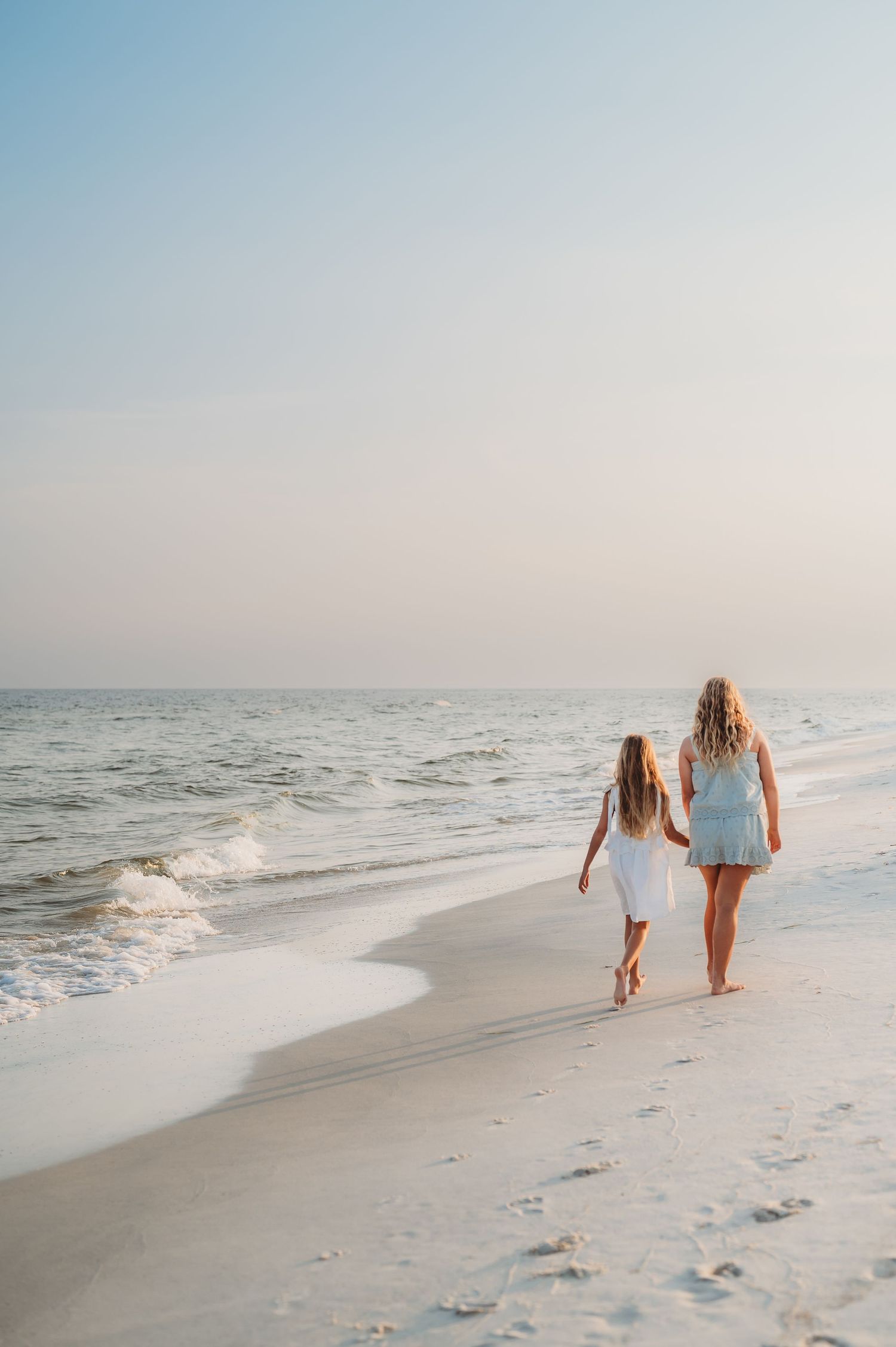 Mother and daughter walking hand in hand on a white sandy beach at sunset wearing white dresses.