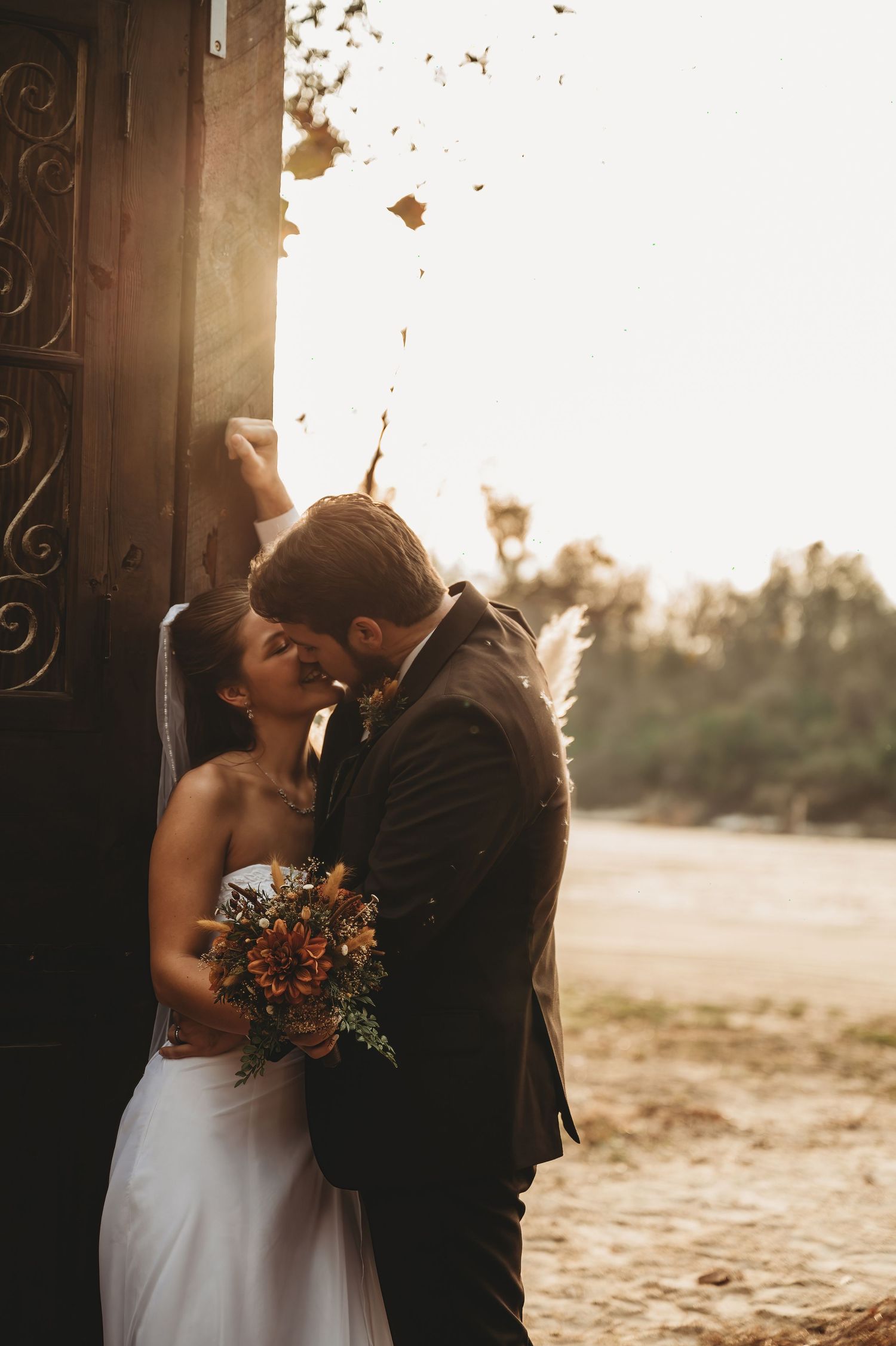 Bride and groom sharing a romantic kiss at golden hour beside an ornate wooden door, bride holding autumn bouquet.