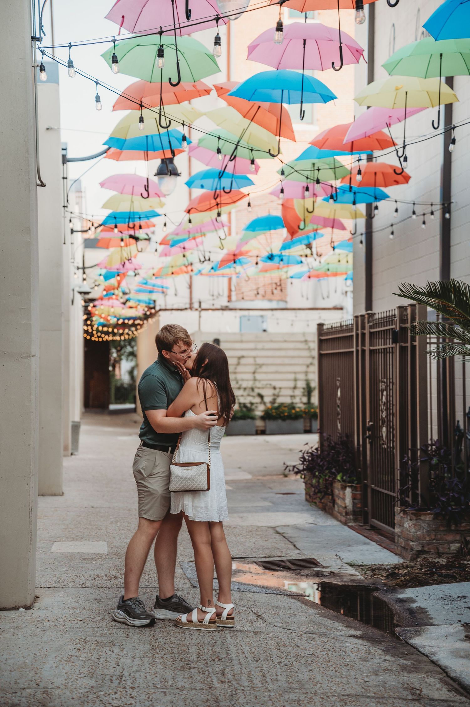 Couple sharing a romantic kiss under colorful hanging umbrellas in a charming outdoor alleyway.