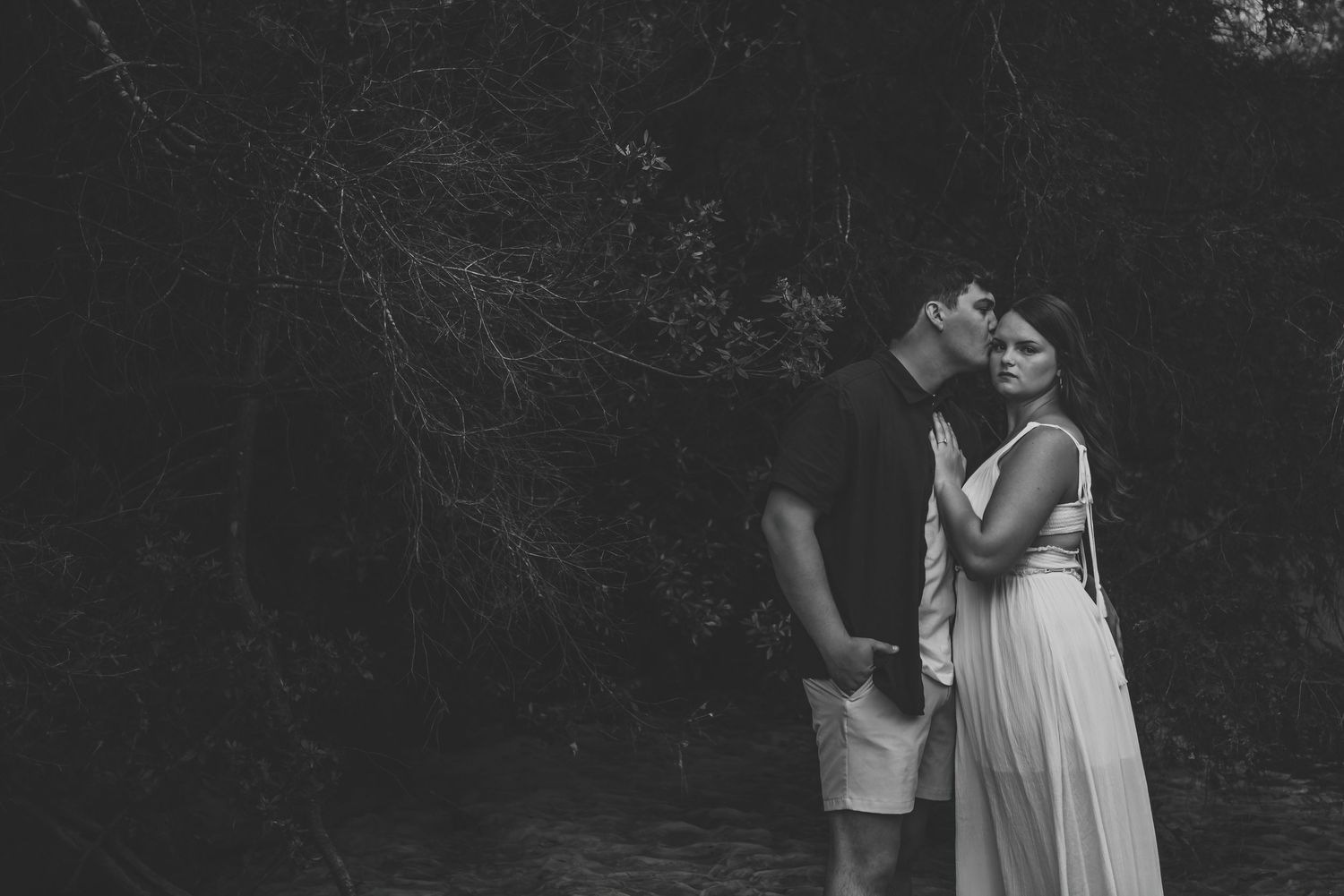 Black and white romantic photo of a couple kissing near a serene woodland stream.