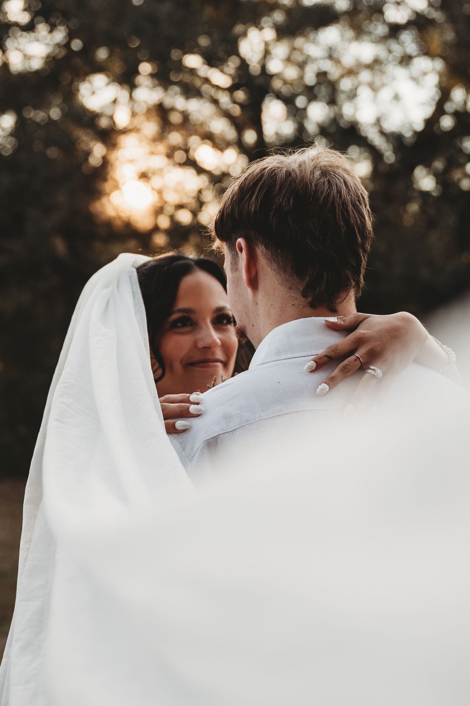 Bride and groom embracing outdoors at golden hour, bride smiling under white veil with bokeh tree background.