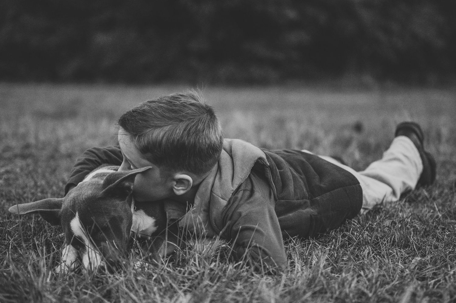 Black and white image of a boy lying on grass affectionately nuzzling a dog in a field.