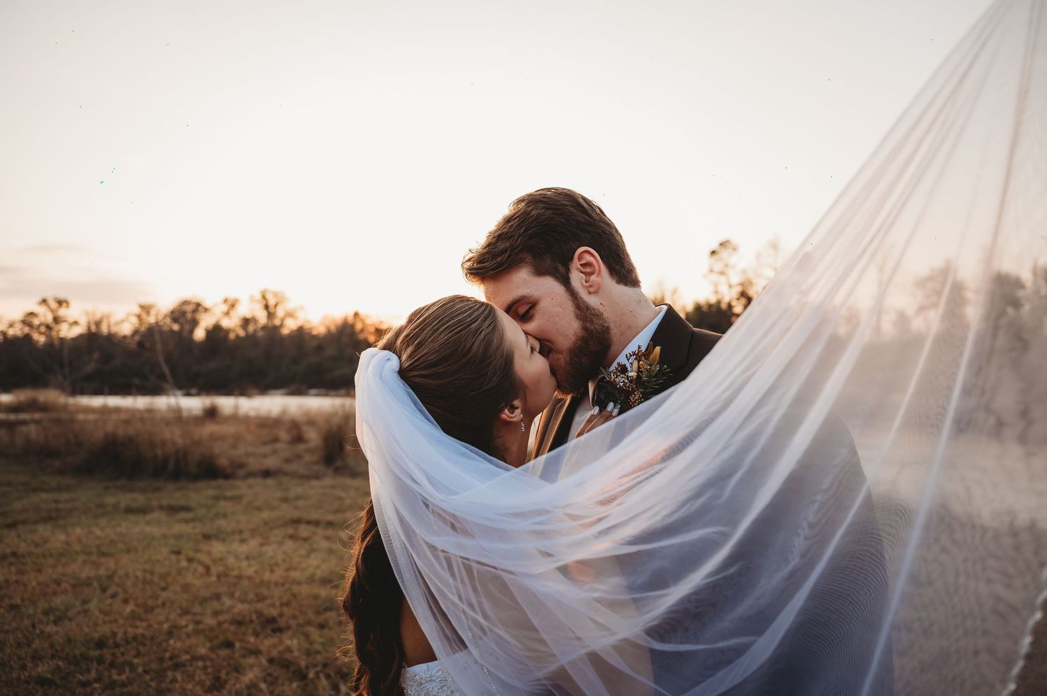 Bride and groom sharing a romantic kiss under a flowing veil at golden hour by a serene lake.