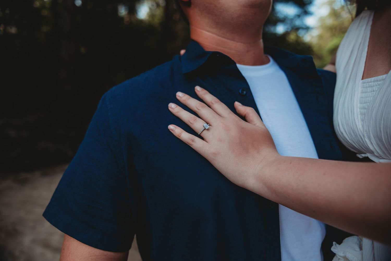 Close-up of a woman's hand with an engagement ring resting on a man's shoulder in a romantic outdoor setting.
