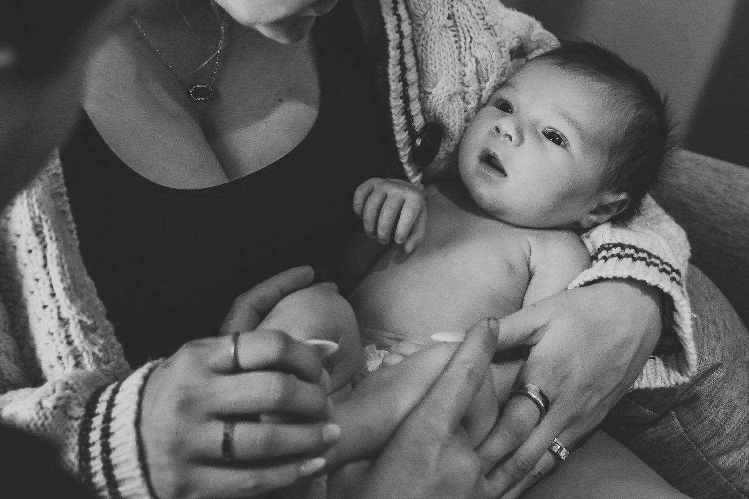 Black and white close-up of mother holding alert newborn baby with fathers hand visible.