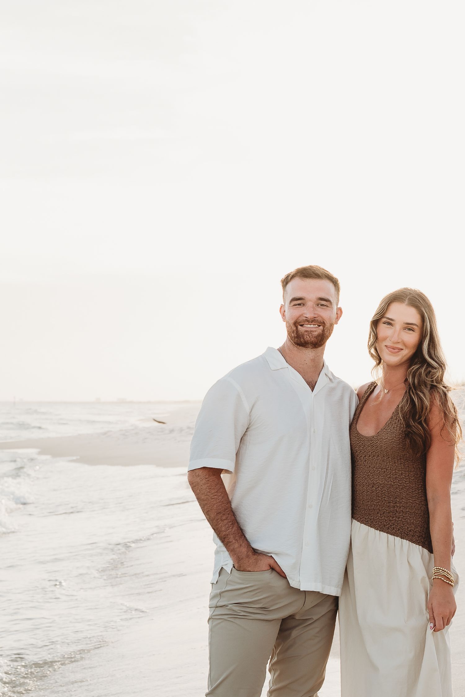 Smiling couple poses together on beach at sunset, man in white shirt and woman in brown top and white pants.