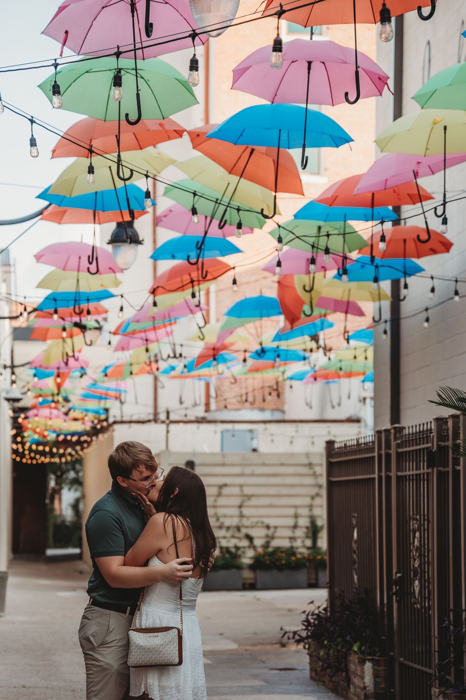 Couple kissing under colorful hanging umbrellas in a vibrant outdoor alley decorated with string lights.