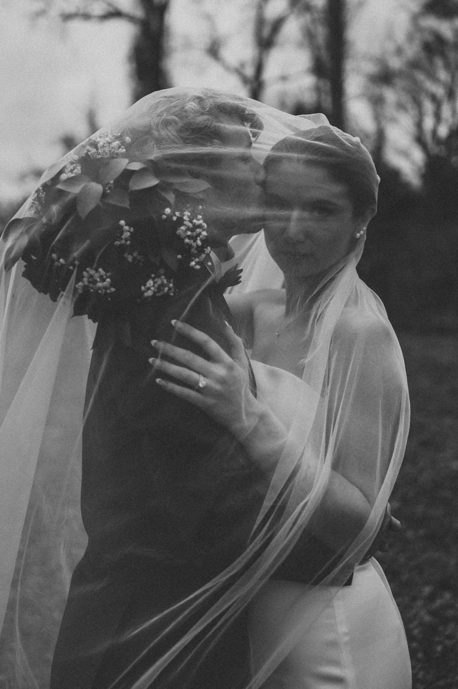 Black and white photo of a bride holding flowers, draped in a flowing veil, embracing her partner outdoors.