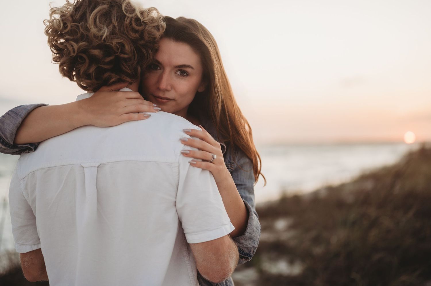 A woman with long brown hair hugs a curly-haired man near the ocean at sunset.