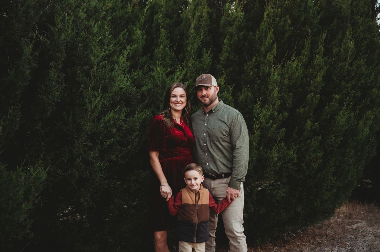 A happy family of three poses outdoors in front of tall green trees during a fall photo session.