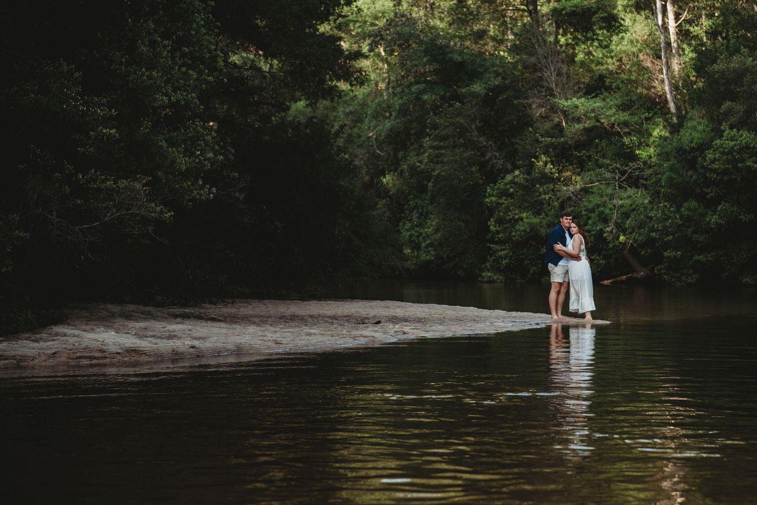 Couple embracing on a sandy riverbank surrounded by dense green forest trees.