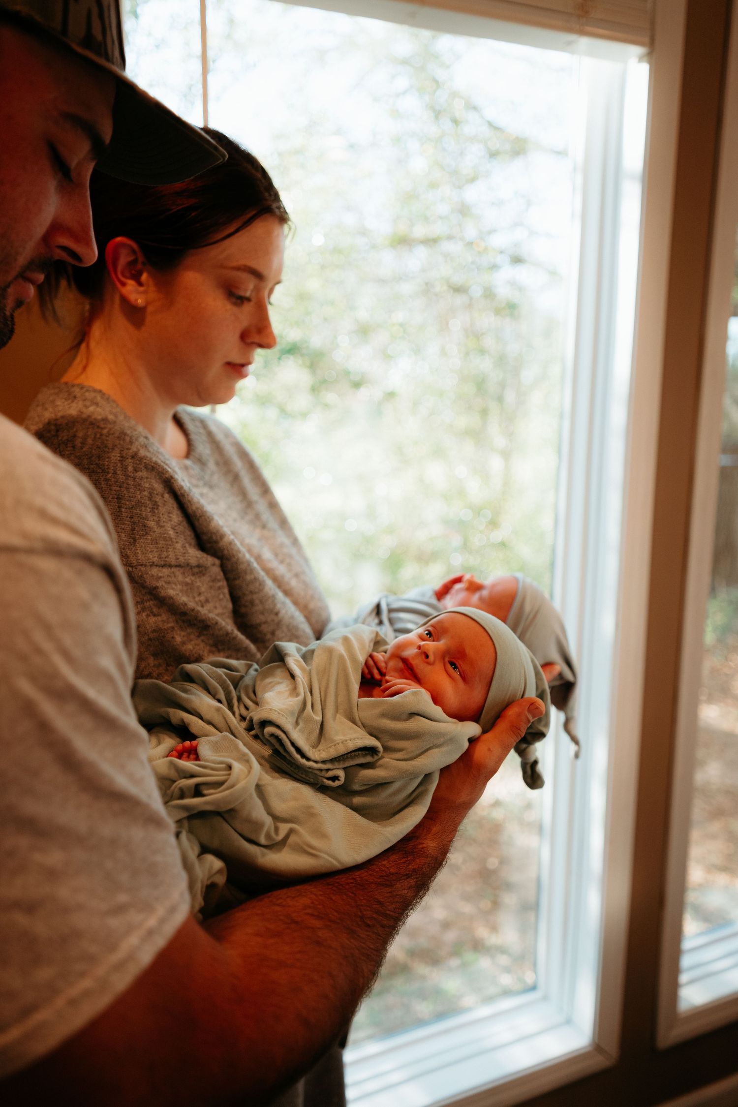 Loving parents gaze down at their sleeping swaddled newborn baby near a bright window together.