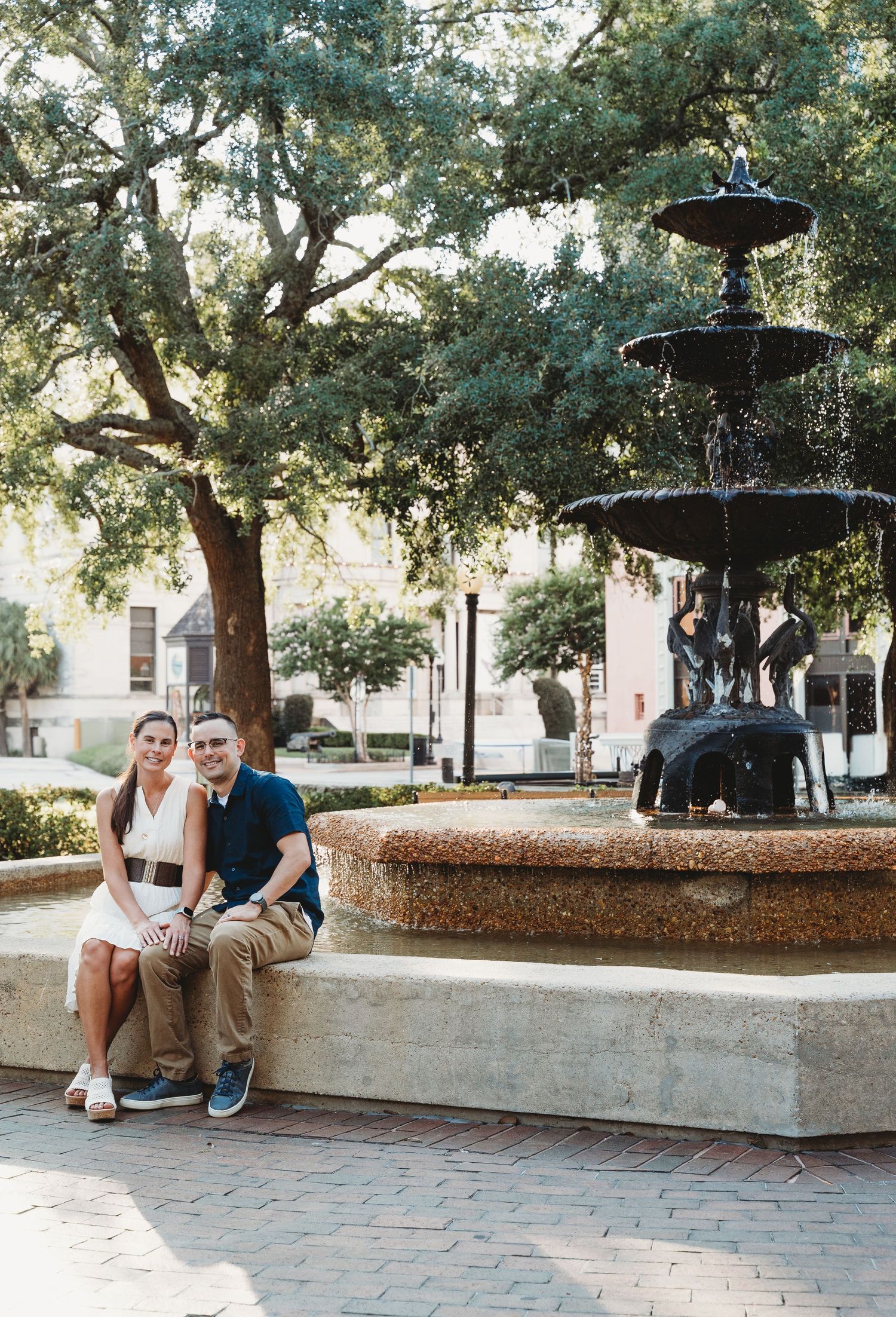 A smiling couple sits together by an ornate tiered fountain beneath a large oak tree.