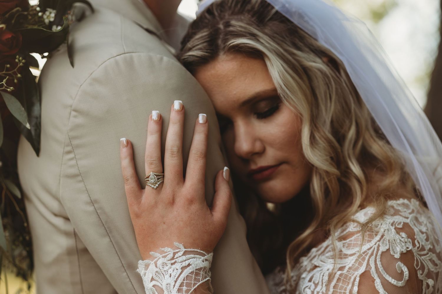 Close-up of bride in lace gown resting head on groom showing elegant diamond ring outdoors.