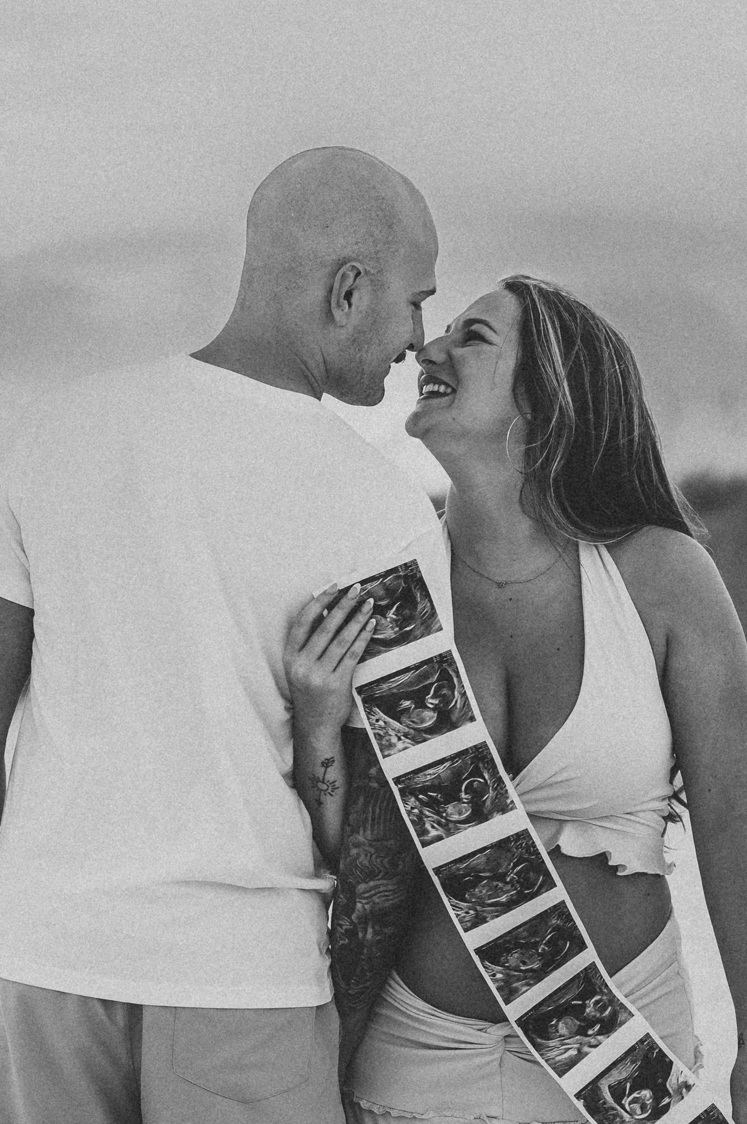 Smiling pregnant couple holding ultrasound strip photos during black and white beach maternity shoot.
