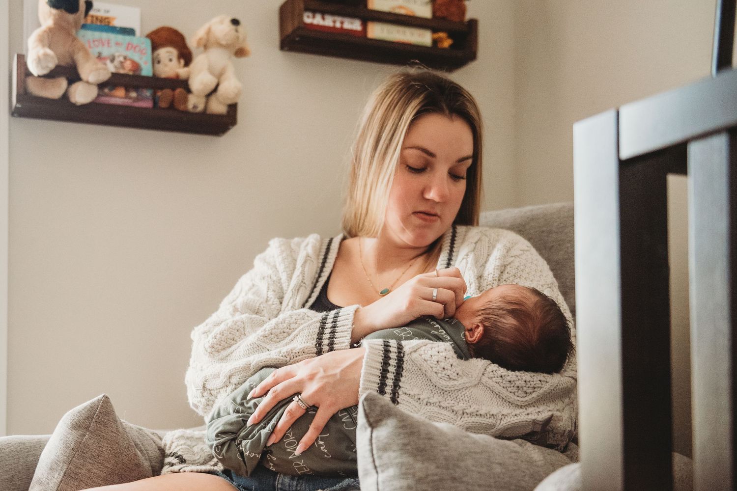 Young mother sitting in gray chair tenderly cradling and nursing her newborn baby.