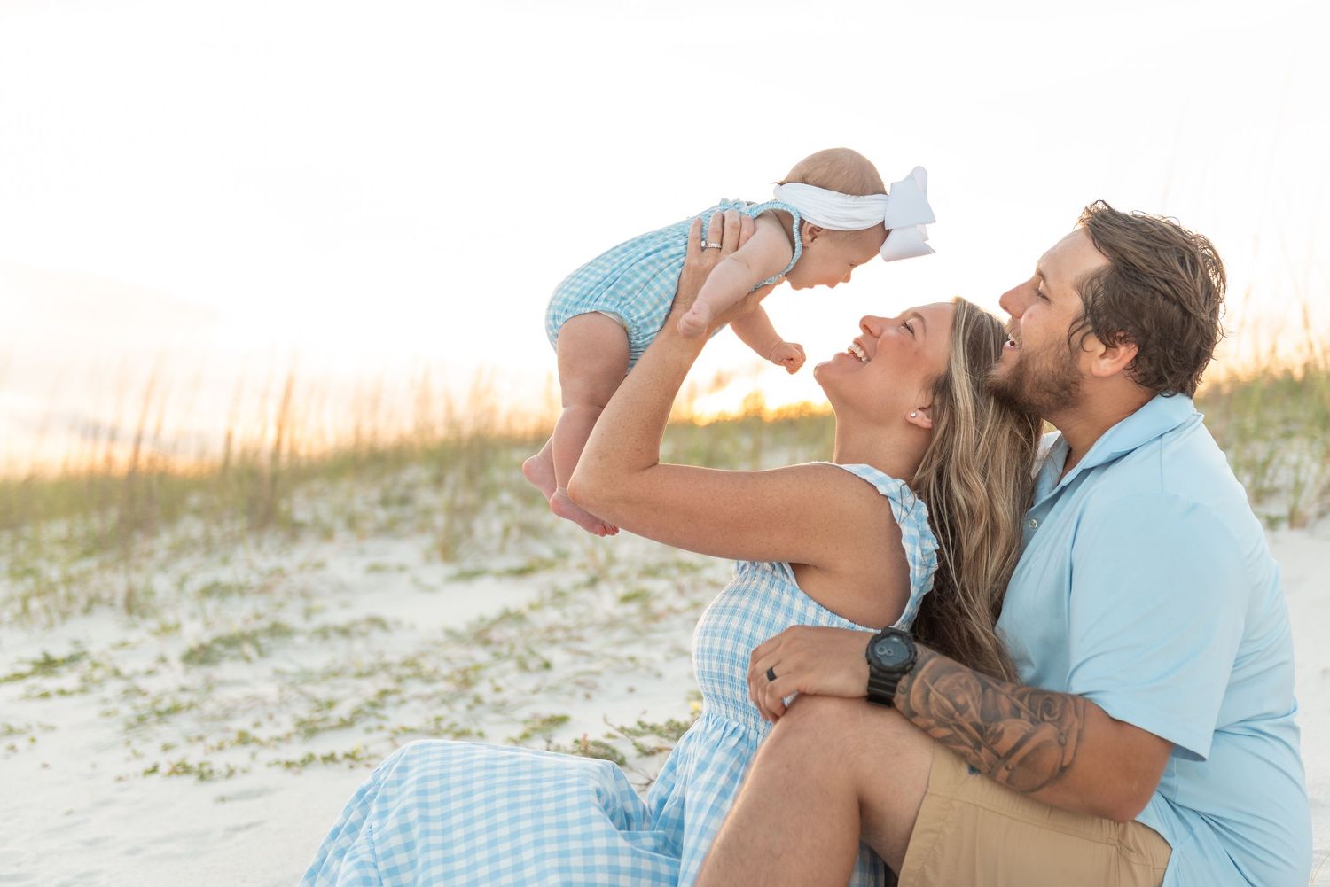Joyful family sitting on beach sand with mom lifting smiling baby toward dad.