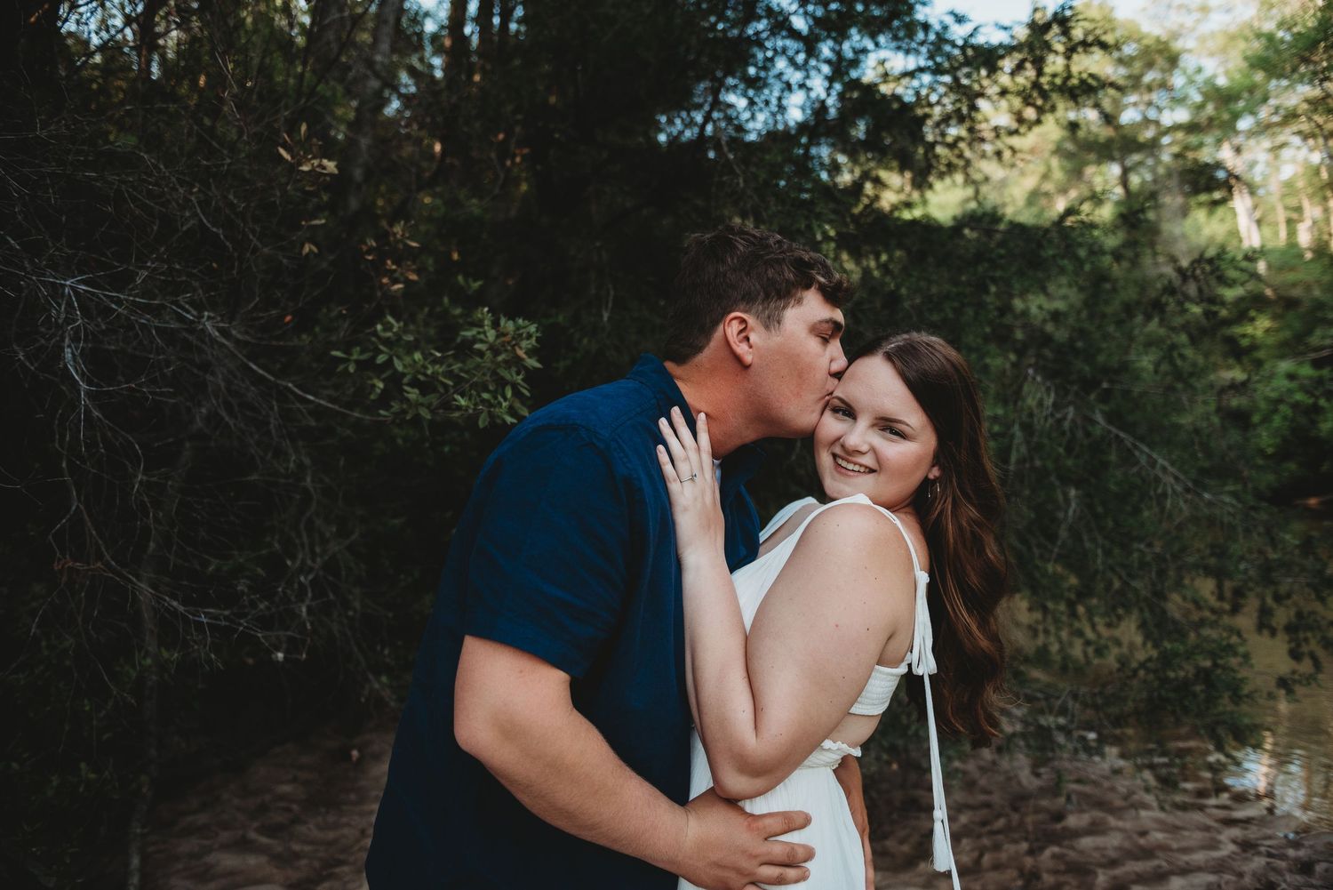 Man kissing woman on the cheek as she smiles near a sunlit river with green trees.