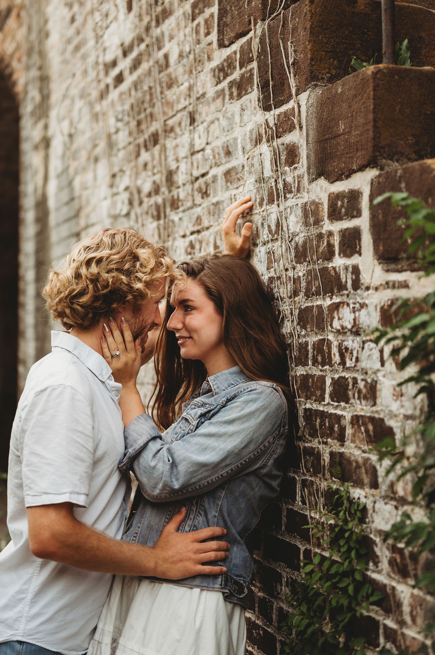 Romantic couple embracing against rustic brick wall, woman in denim jacket leaning back as man leans in close.