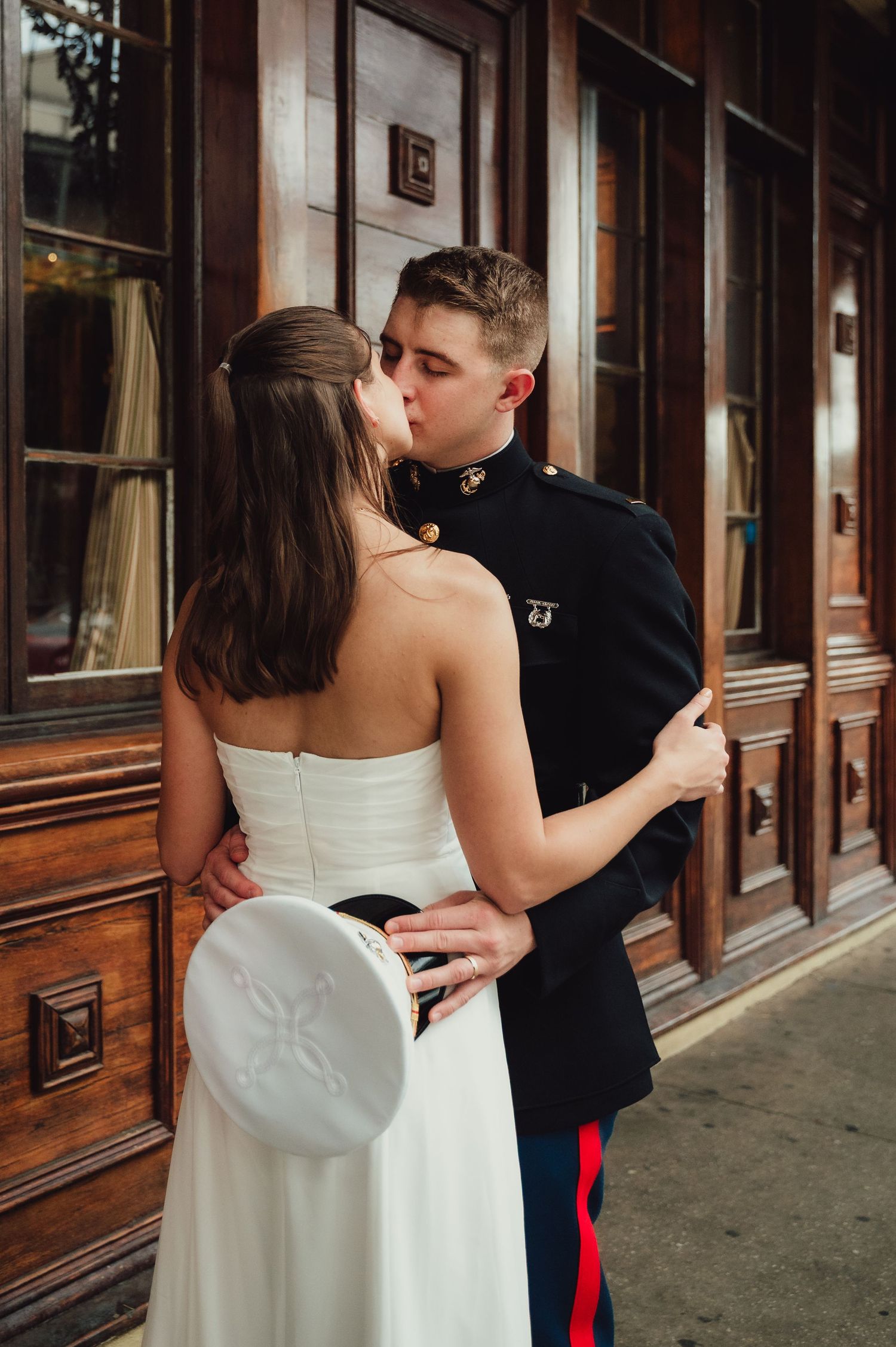 Marine in dress uniform kissing bride in white strapless gown outside a wooden building.