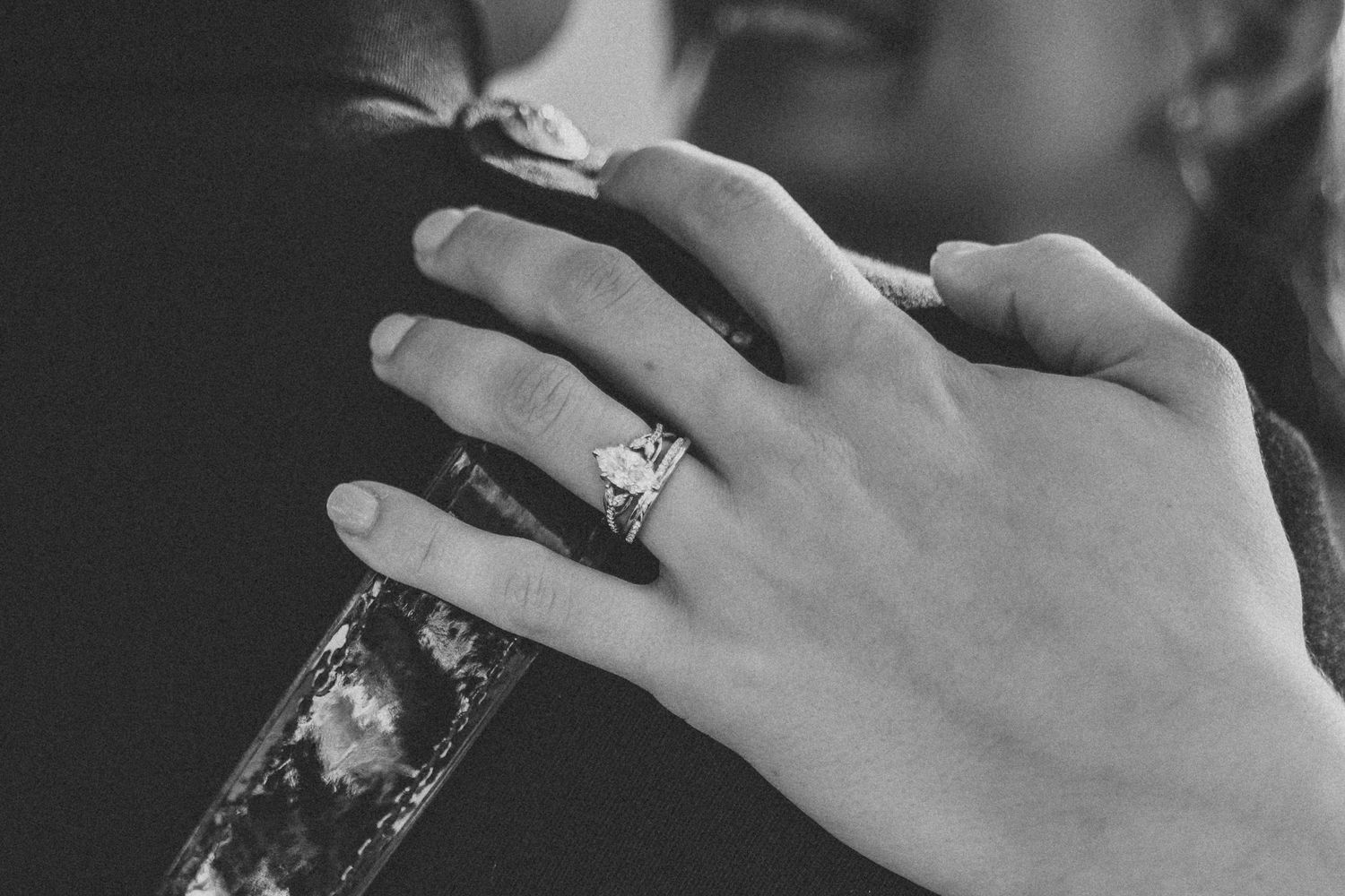 Close-up black and white photo of a woman's hand wearing a diamond engagement ring, resting on a dark surface.