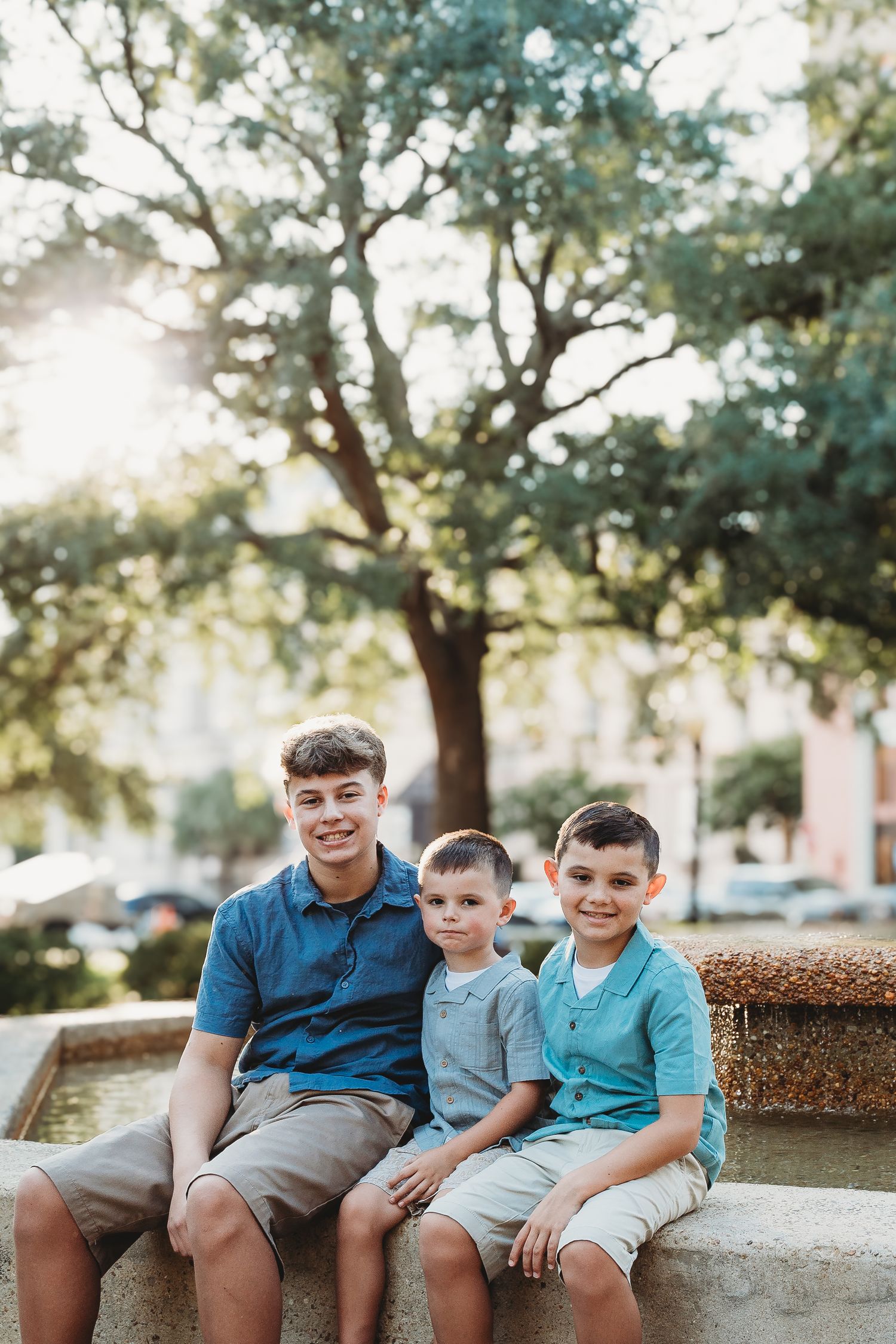 Three brothers smile together while seated on a fountain ledge beneath a large oak tree.