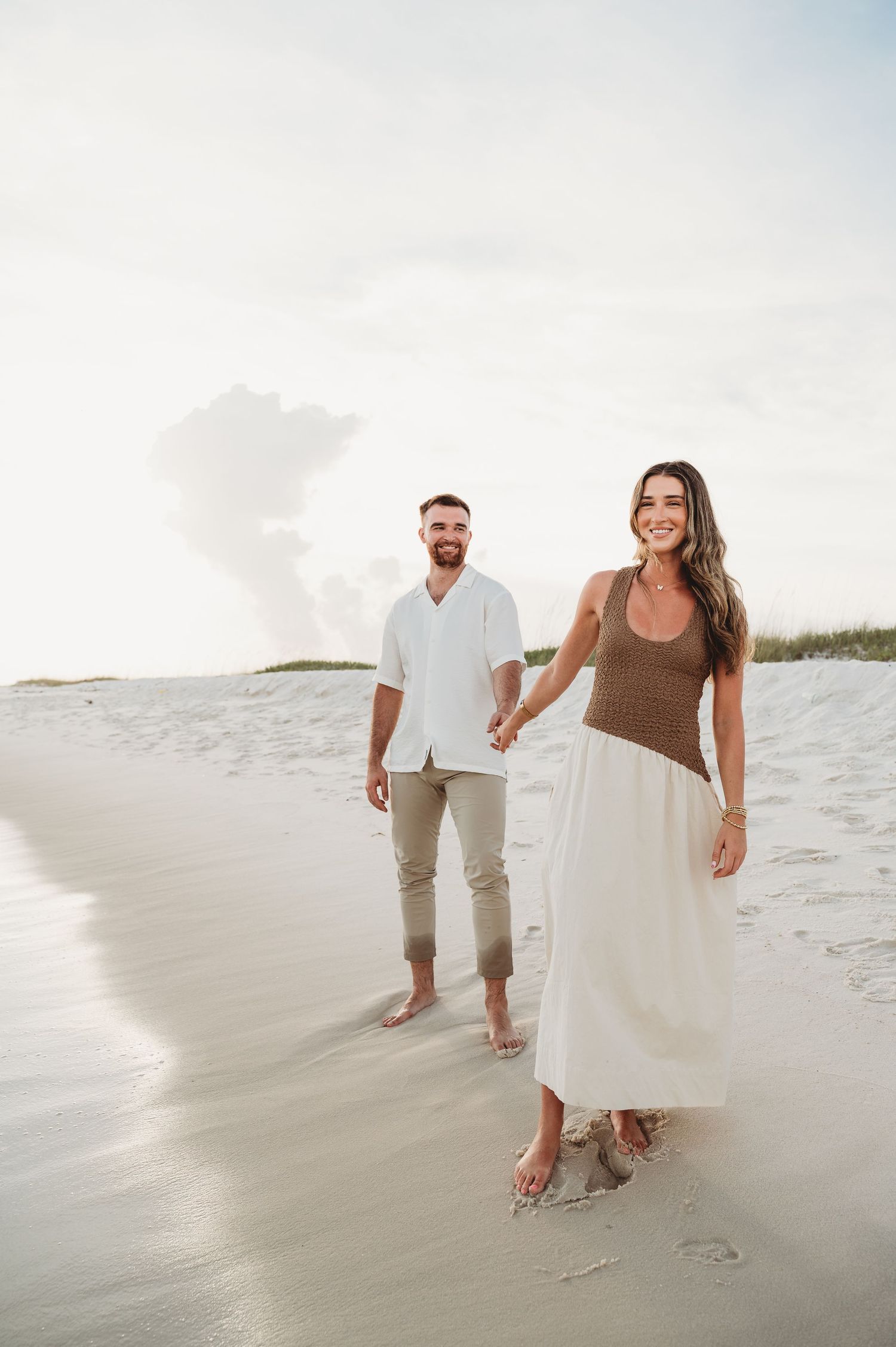 Couple walking on white sand beach, woman in brown top and white skirt leads man in white button-down.