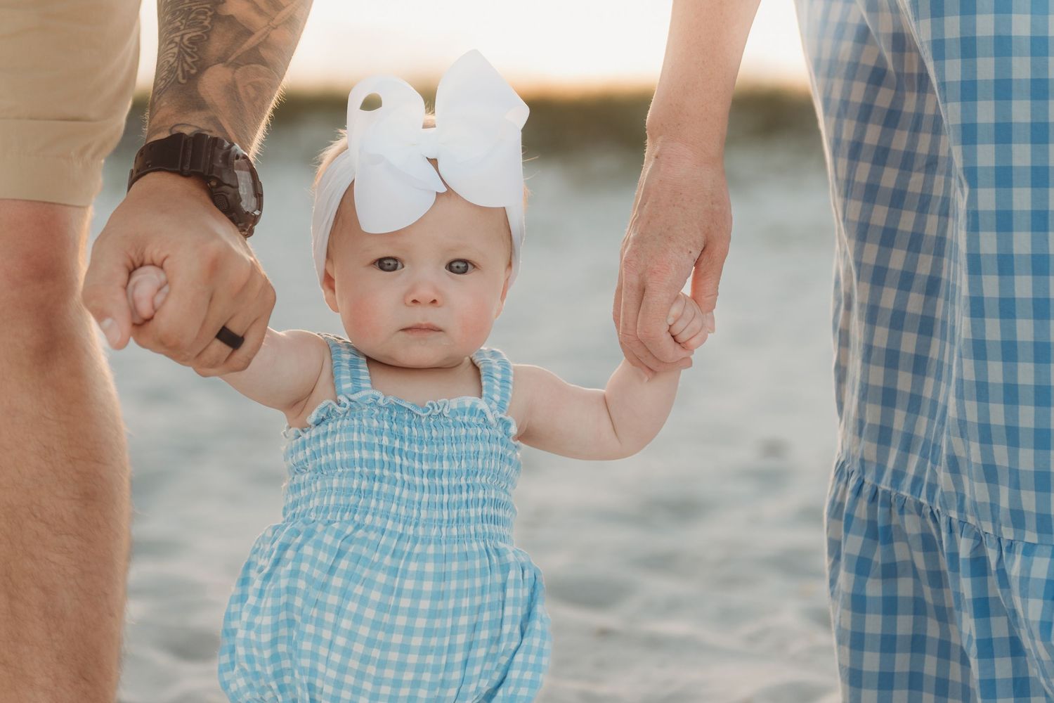 Close-up of baby girl in blue gingham outfit holding both parents hands on beach.