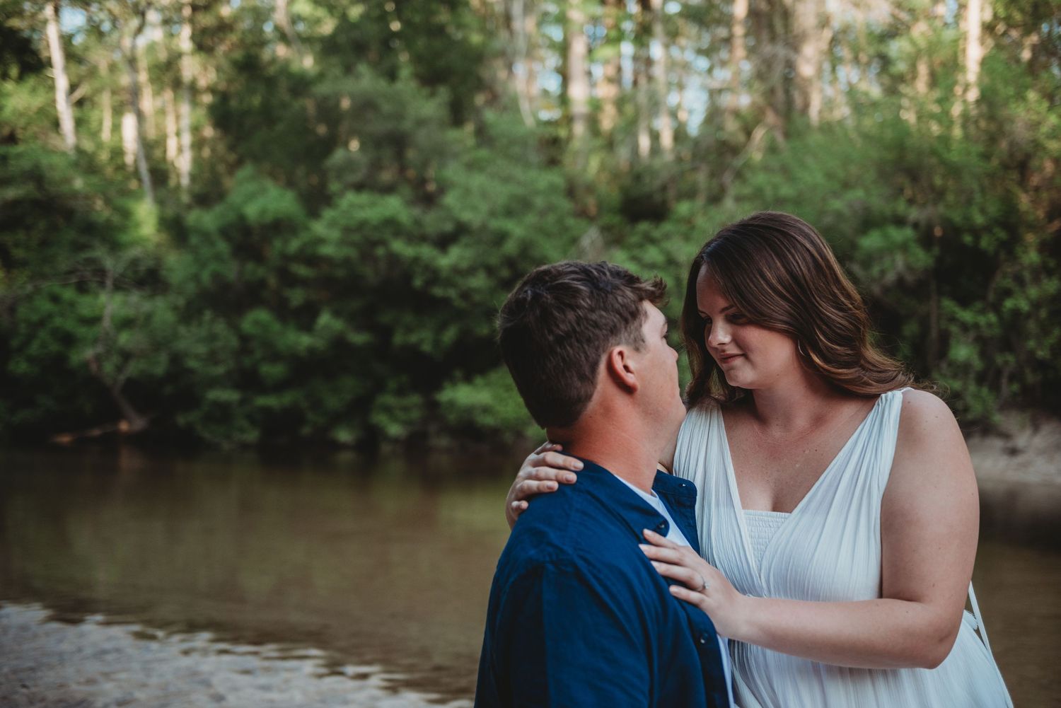 Smiling couple embracing near a peaceful river with dense green trees in the background.