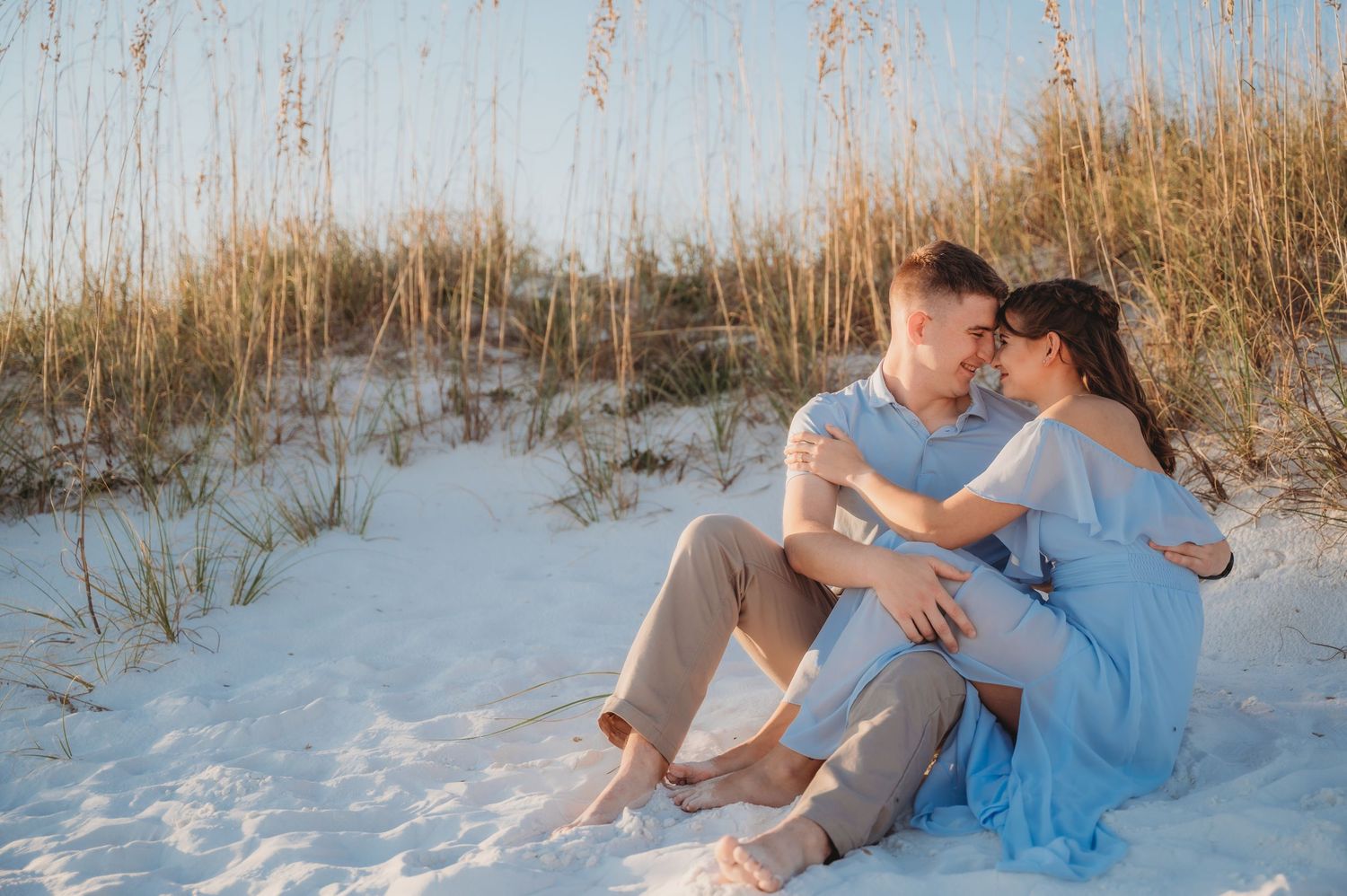 Couple sitting together on white sand dunes at sunset, woman in blue dress, man in khaki, sharing a romantic moment.