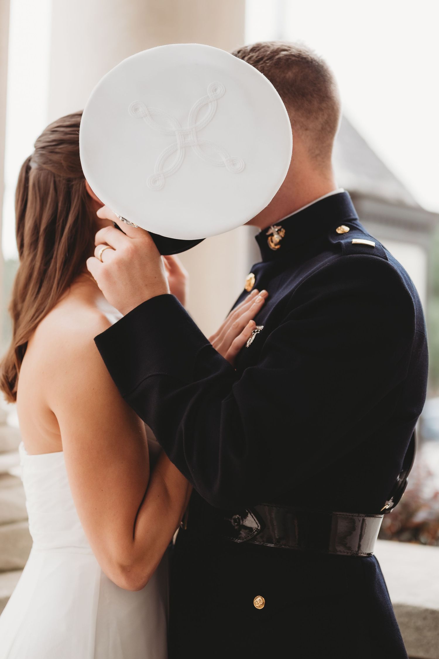 Marine in dress uniform kissing bride, hiding kiss behind white military cover hat on wedding day.