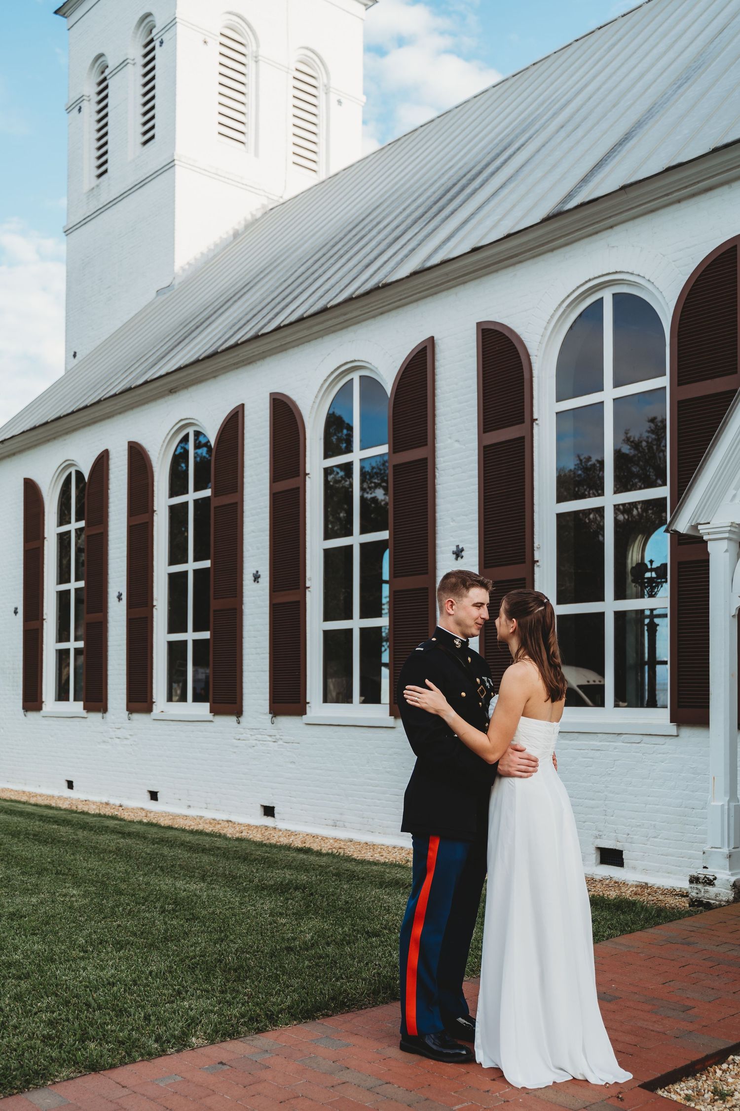 Military groom in dress blues holds bride in white gown outside historic white church.